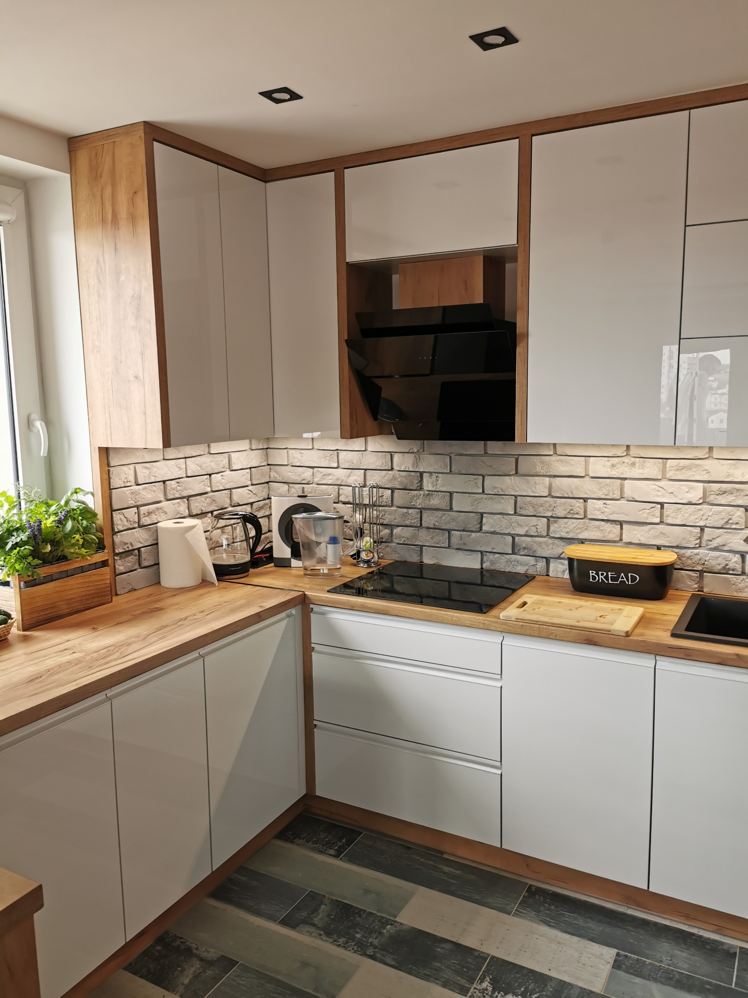 Nowoczesna kuchnia z białymi frontami szafek, drewnianymi blatami i obudową, ceglaną ścianą nad blatem oraz czarną płytą indukcyjną i pochłaniaczem.