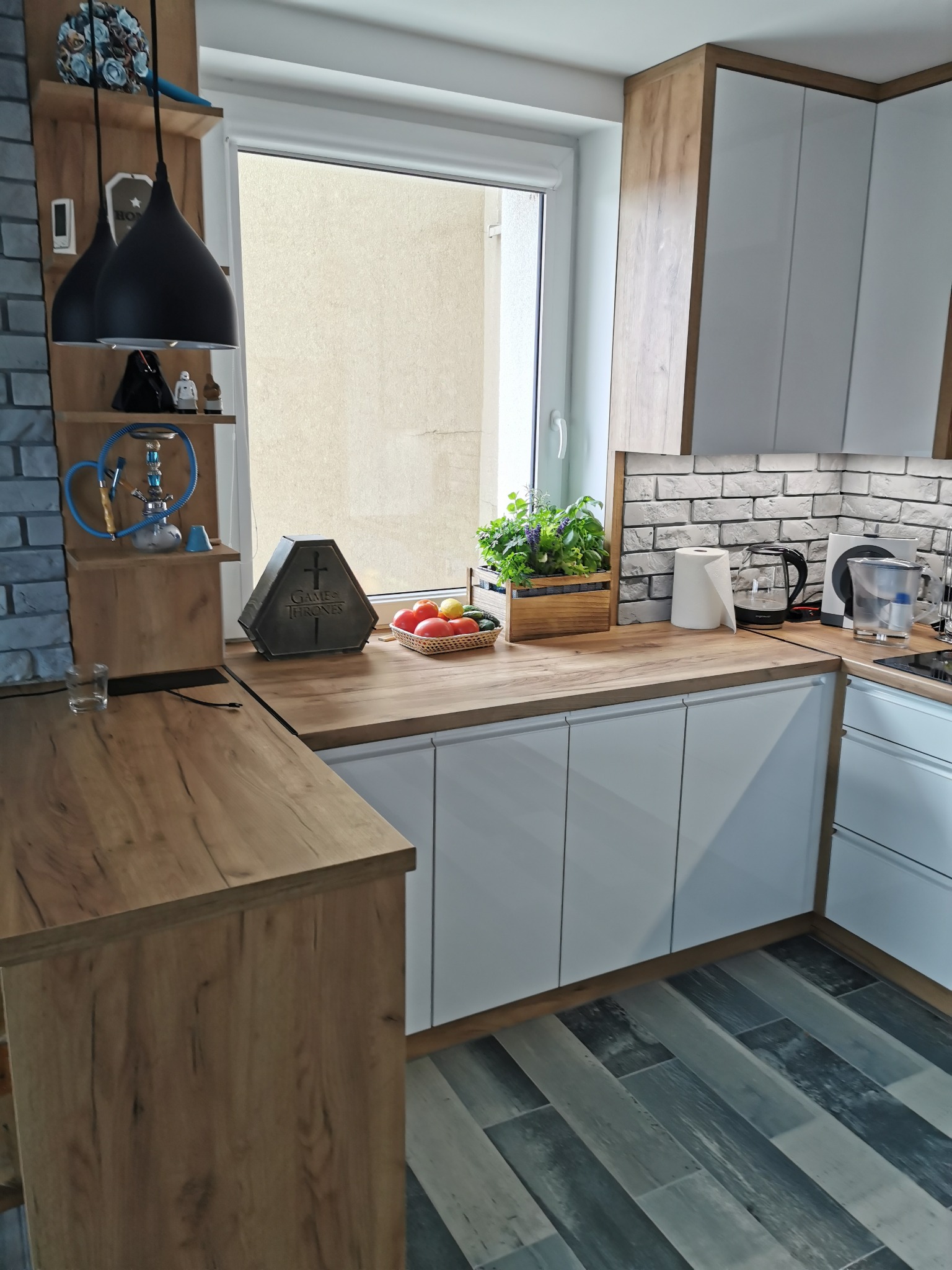 Nowoczesna kuchnia z białymi frontami szafek i drewnianym blatem, uzupełniona o dekoracje takie jak lampy wiszące, rośliny w drewnianej skrzynce i ozdobny pojemnik z motywem Gry o Tron.