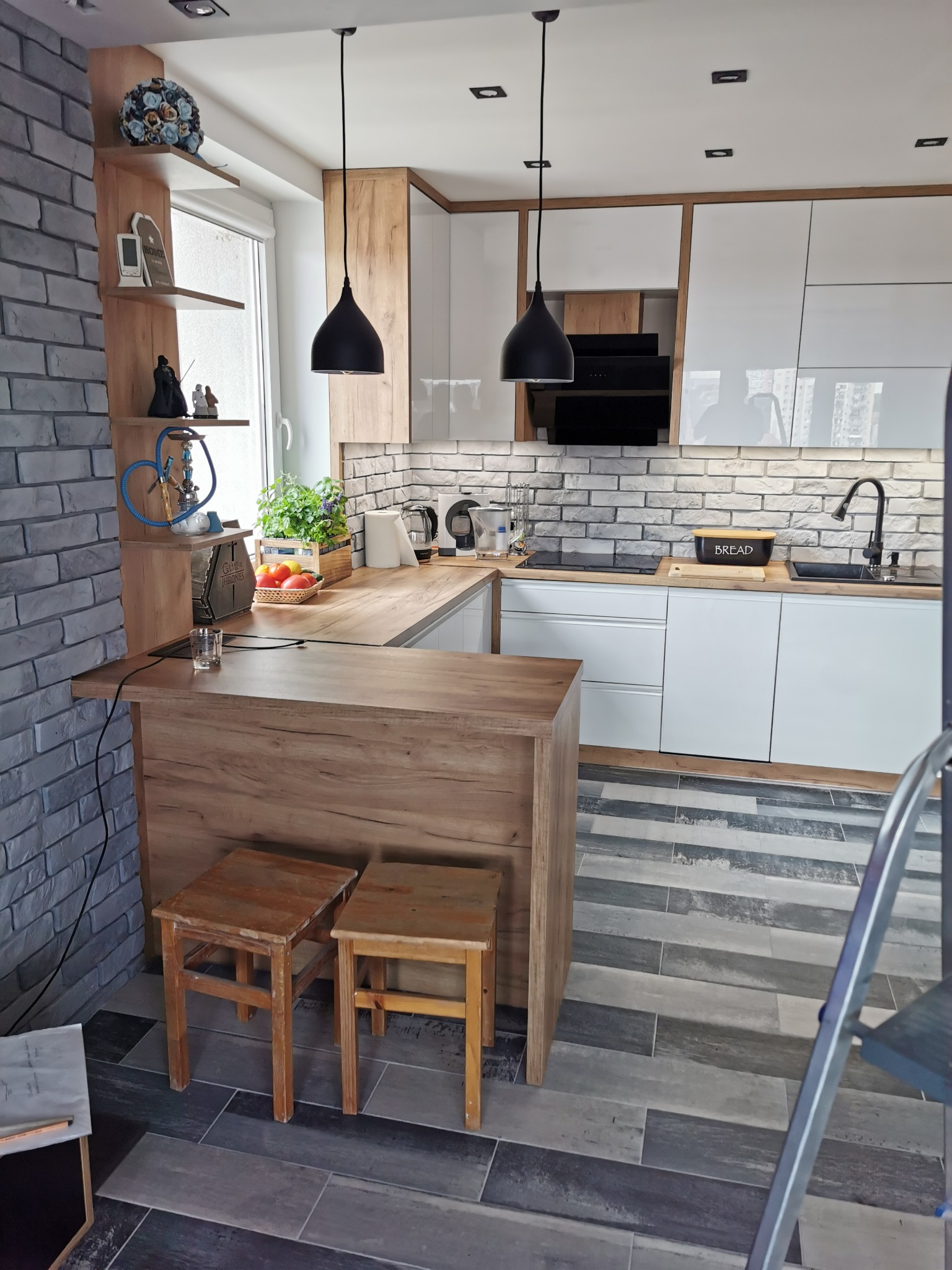 Nowoczesna kuchnia z białymi szafkami, drewnianym blatem i barkiem, oświetlona czarnymi lampami wiszącymi, z akcentem ceglanej ściany i drewnianych stołków barowych.