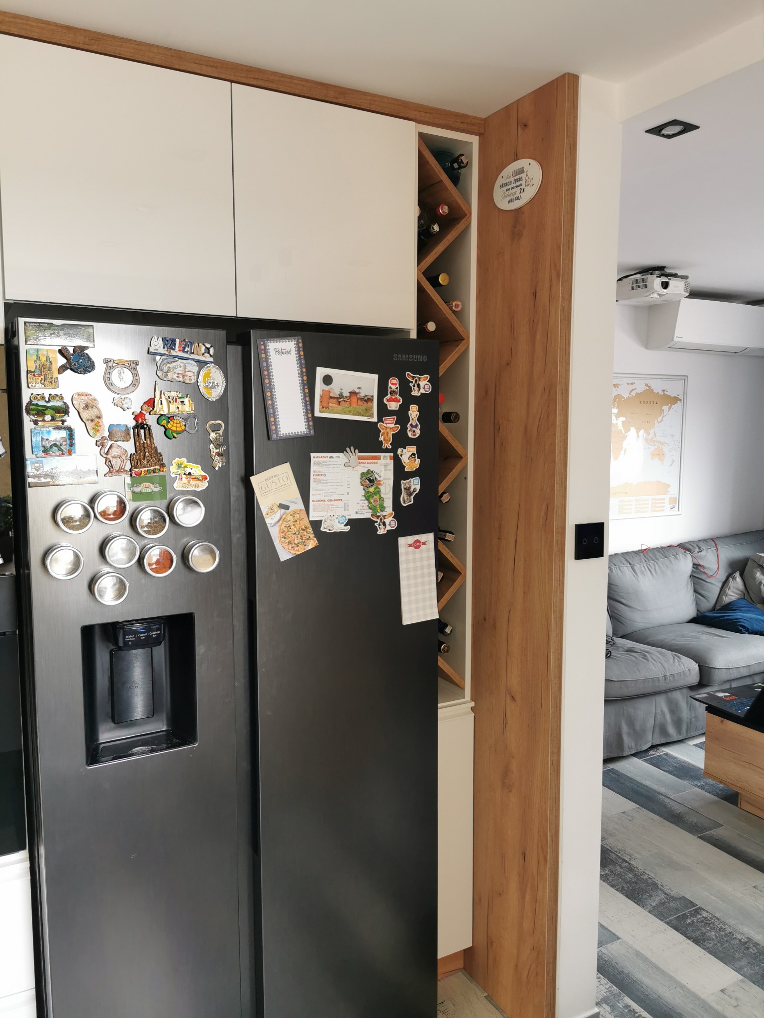 Nowoczesna kuchnia z zabudową szafek, czarną lodówką ozdobioną magnesami i drewnianym regałem na wino w zabudowie.