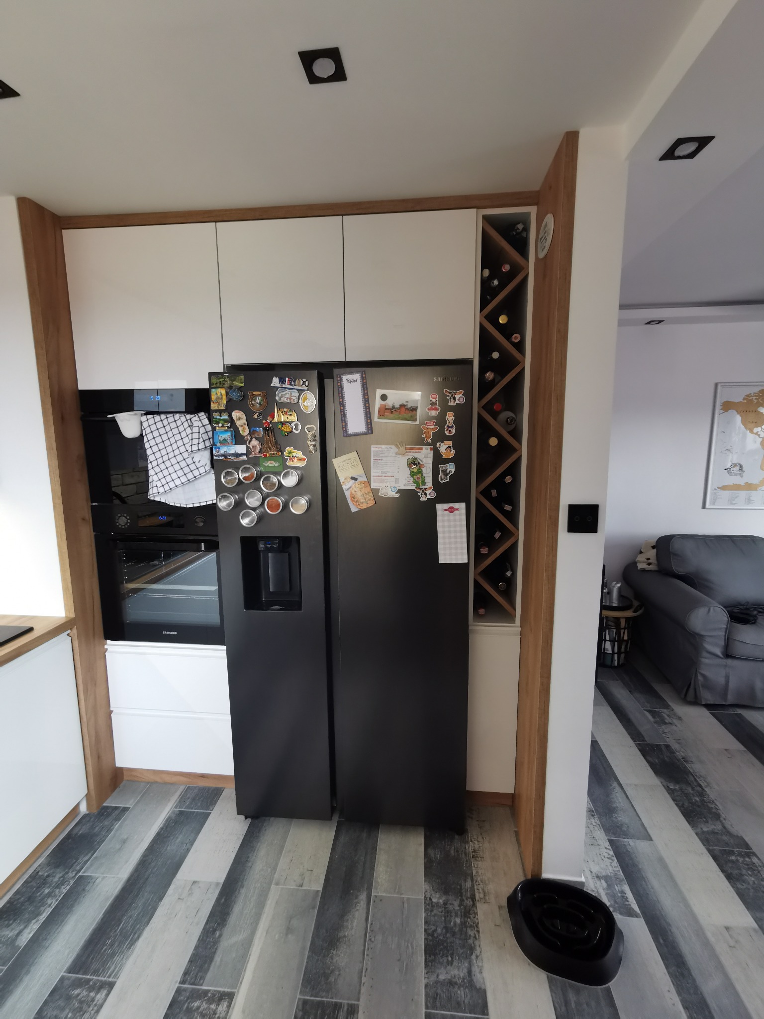 Nowoczesna kuchnia z białymi szafkami, czarną lodówką z magnesami i dozownikiem wody, piekarnikiem, drewnianym regałem na wino oraz szaro-białą podłogą z paneli.