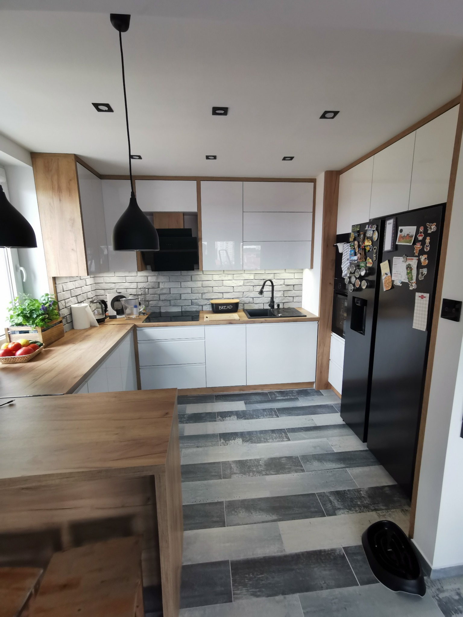 Nowoczesna kuchnia z białymi szafkami, drewnianymi akcentami i szarą podłogą w paski. Czarny zlew i lodówka z magnesami. Na blacie koszyk z owocami i zioła w doniczce.