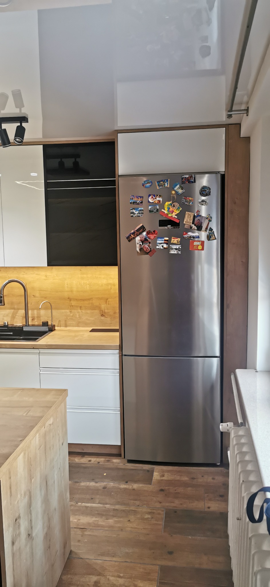 Nowoczesna kuchnia z białymi szafkami, drewnianymi blatami, czarnymi akcentami i lodówką obłożoną magnesami, widok z perspektywy podłogi z drewnopodobnych paneli.