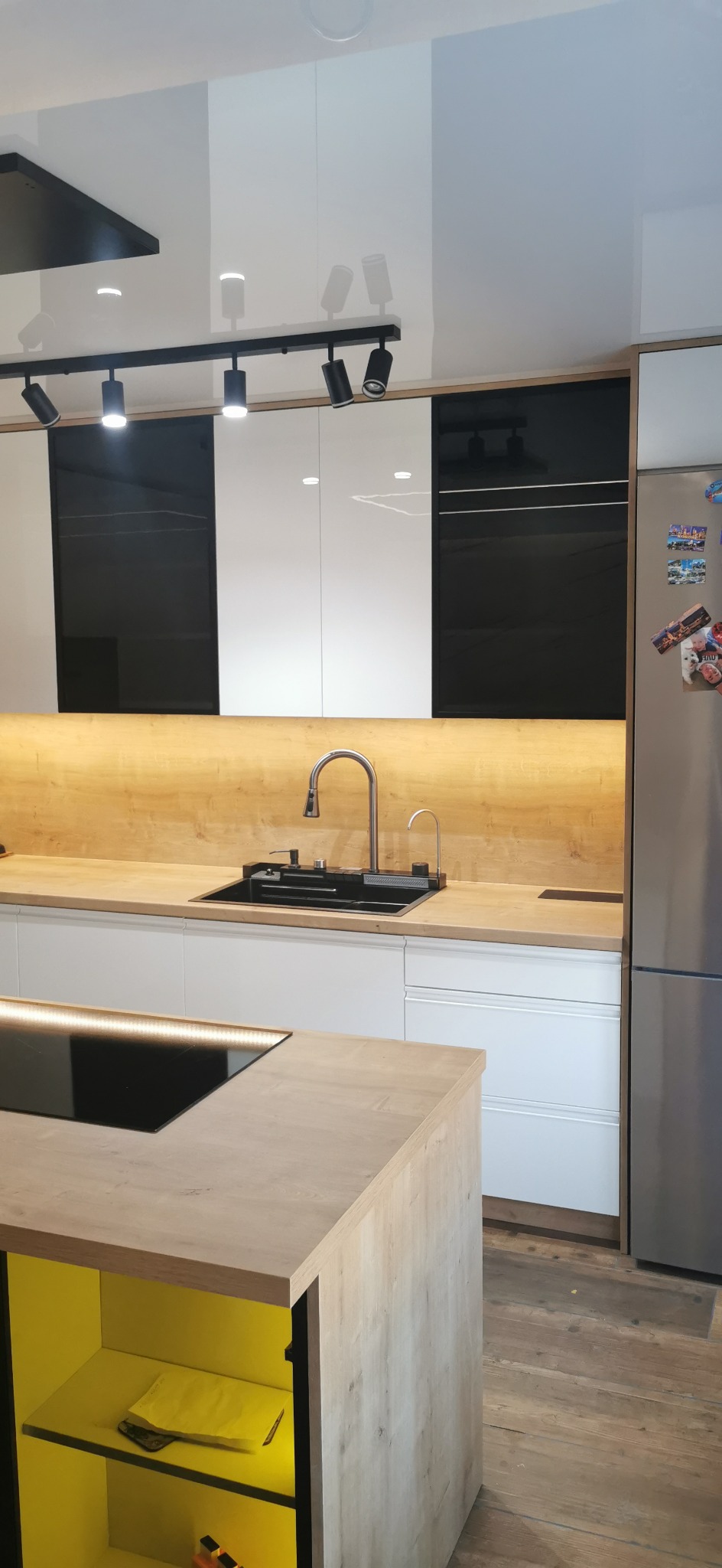 Nowoczesna kuchnia z białymi i czarnymi szafkami, drewnianym blatem i wyspą z żółtym wnętrzem, oświetlona lampami punktowymi.