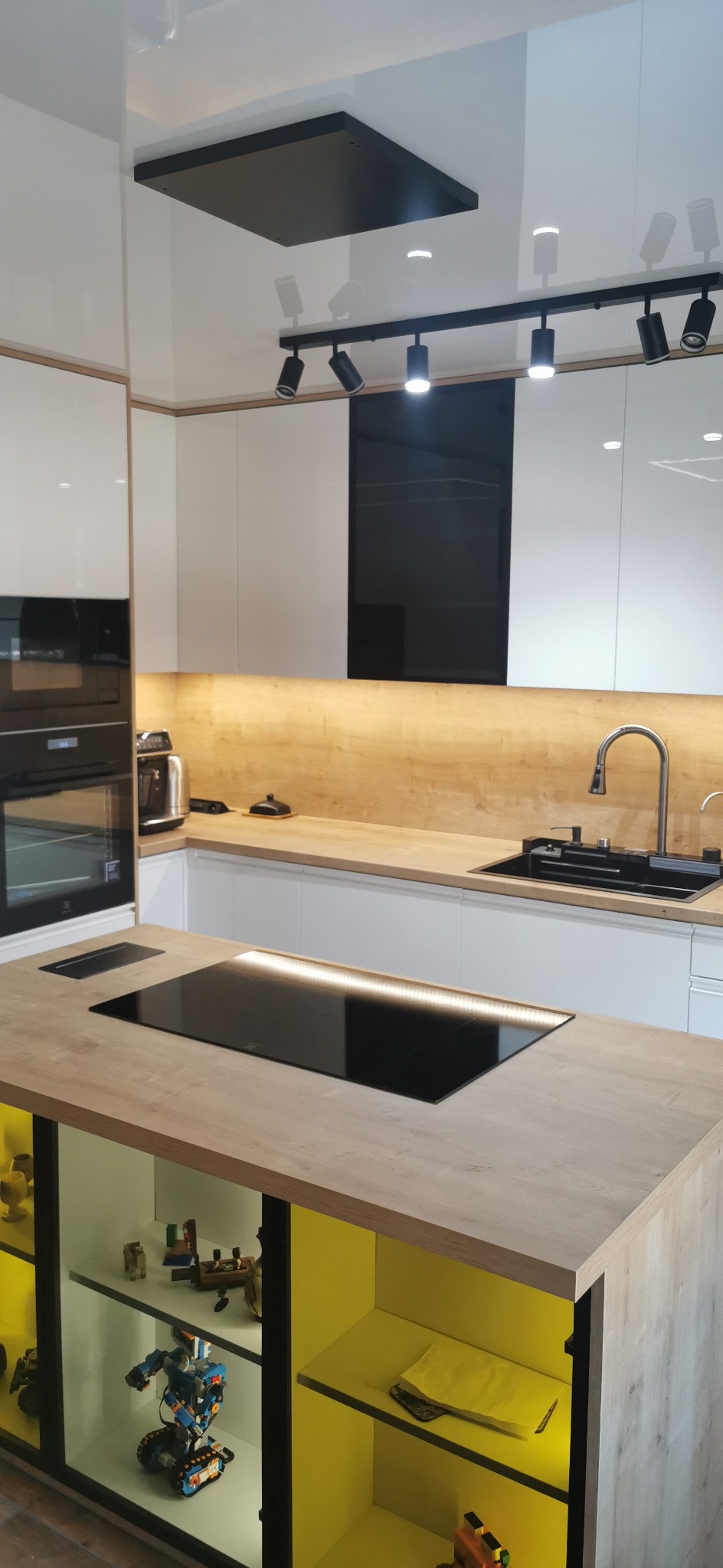 Nowoczesna kuchnia z wyspą: białe, lakierowane szafki, drewniany blat z indukcyjną płytą, czarny zlew z baterią, oświetlenie punktowe. W wyspie przeszklone półki z żółtym podświetleniem...
