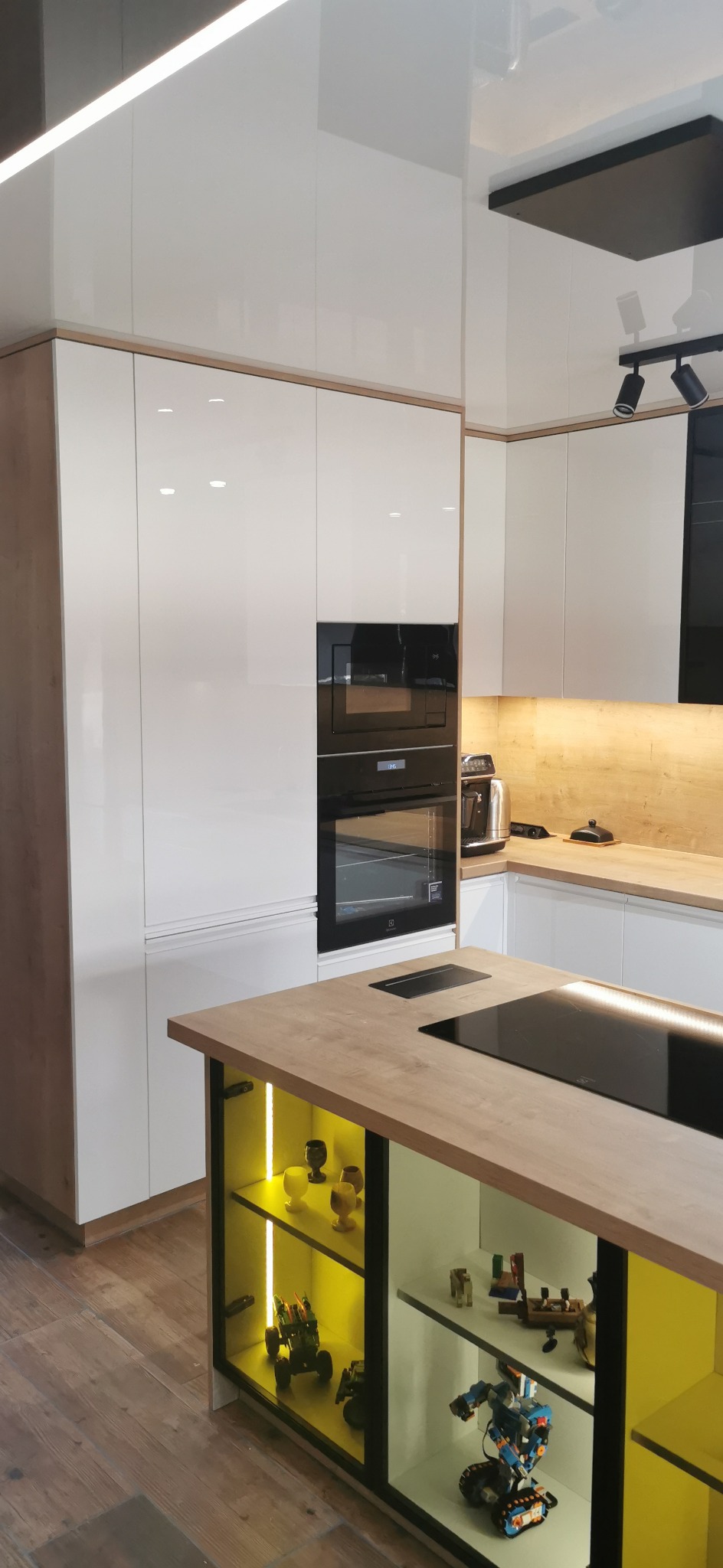 Nowoczesna kuchnia z białymi, lakierowanymi frontami, drewnianym blatem i wyspą z podświetlanymi półkami, eksponującymi kolekcję figurek.