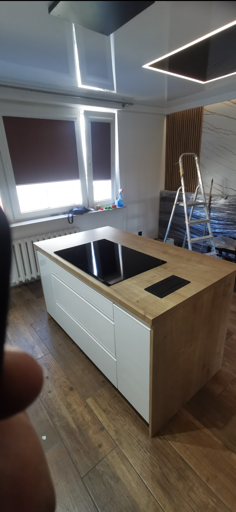 Wyspa kuchenna z drewnianym blatem i białymi frontami szuflad, wbudowana płyta indukcyjna, widoczna drabina w tle, pomieszczenie w trakcie wykańczania.