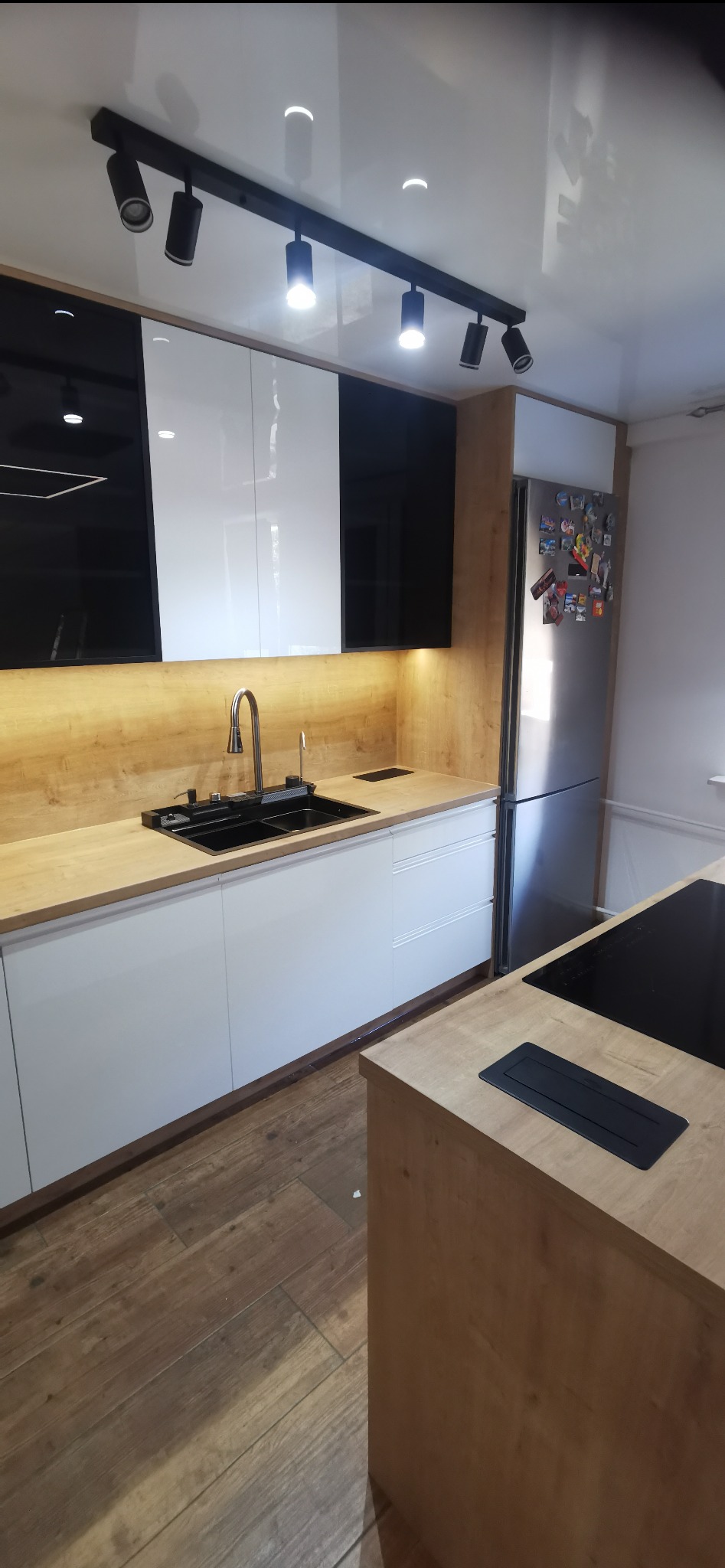 Nowoczesna kuchnia z białymi i czarnymi szafkami górnymi, drewnianym blatem, stalowym zlewem i wyspą z płytą indukcyjną.