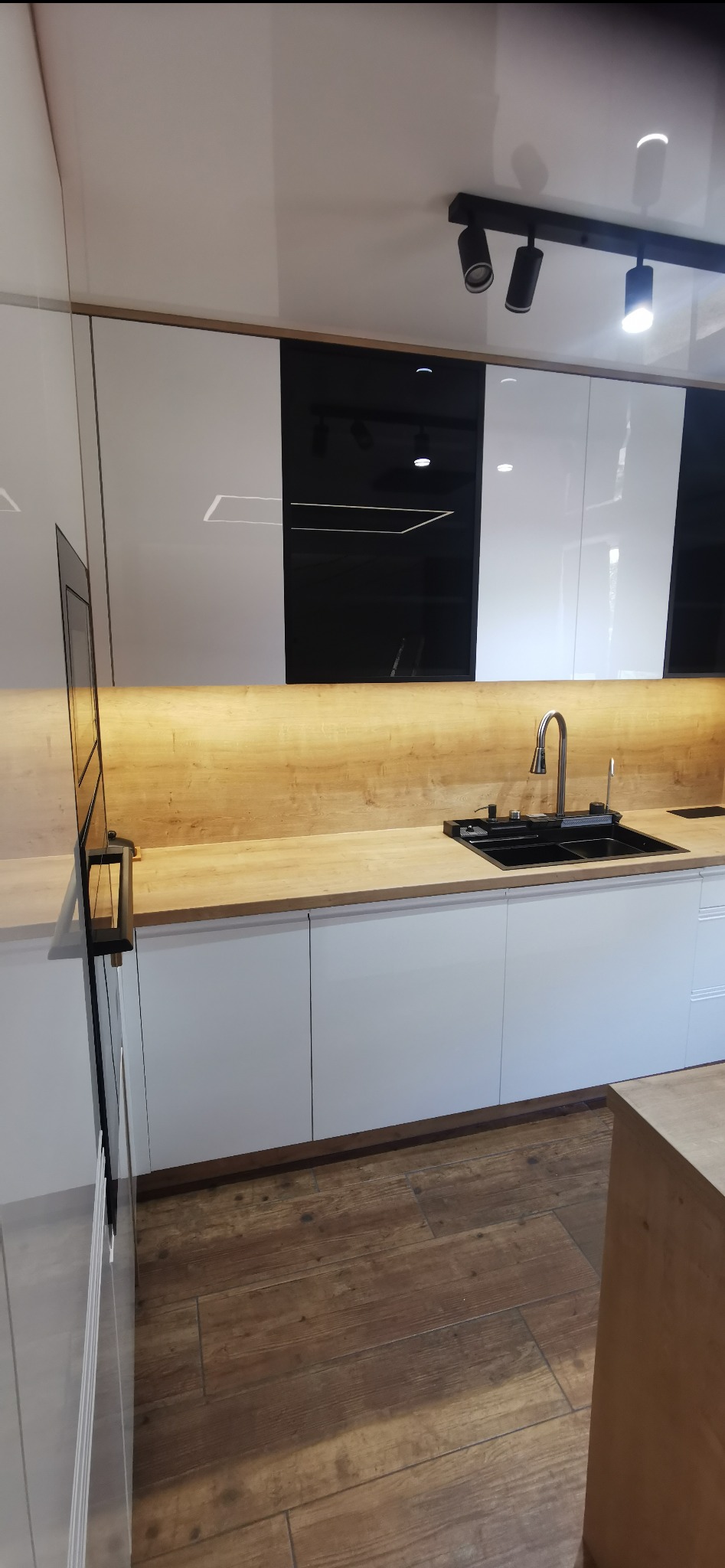 Nowoczesna kuchnia z białymi frontami szafek, drewnianym blatem i czarnymi akcentami, oświetlona punktowo od góry.
