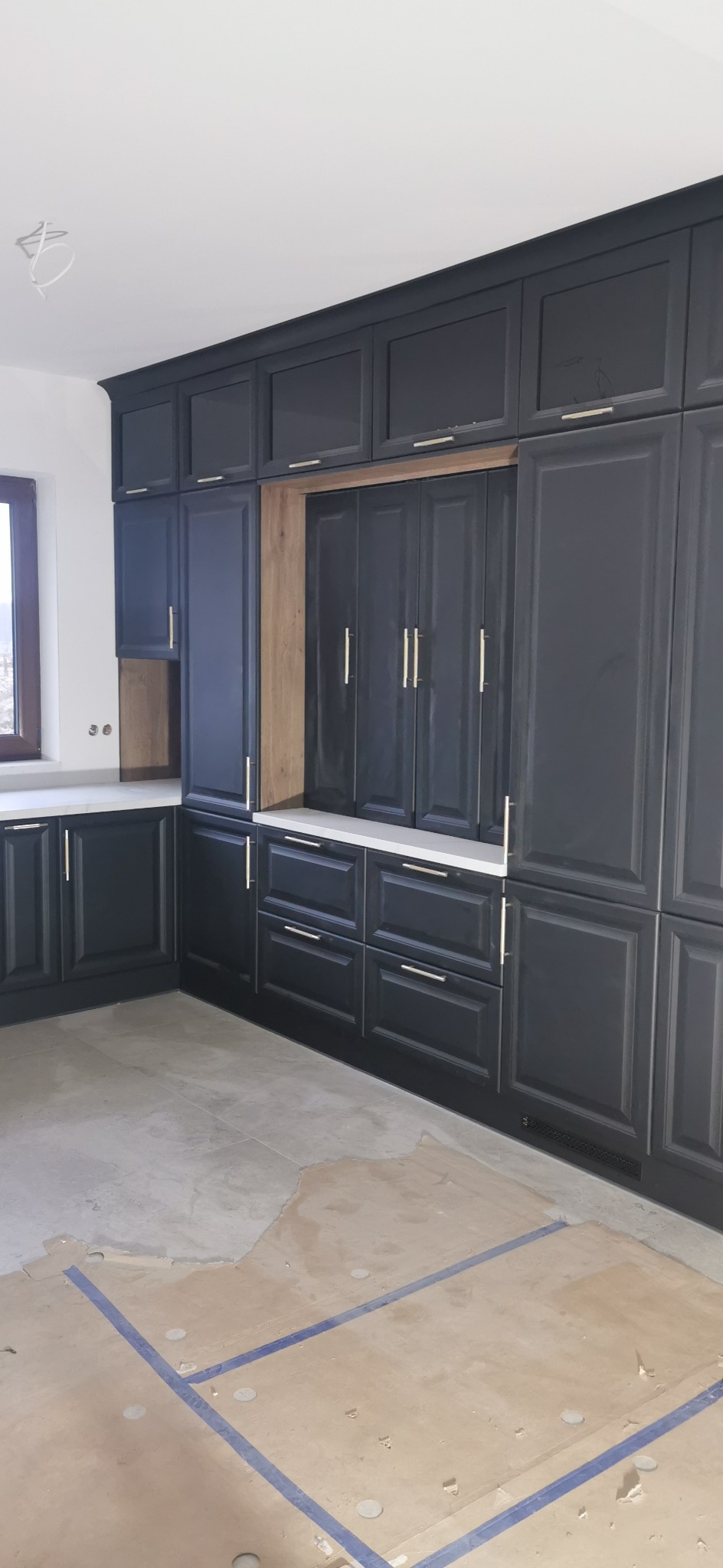 Ciemnoszara zabudowa kuchenna z górnymi szafkami pod sufit, z drewnianymi wstawkami i złotymi uchwytami, na podłodze widoczne ślady po remoncie.