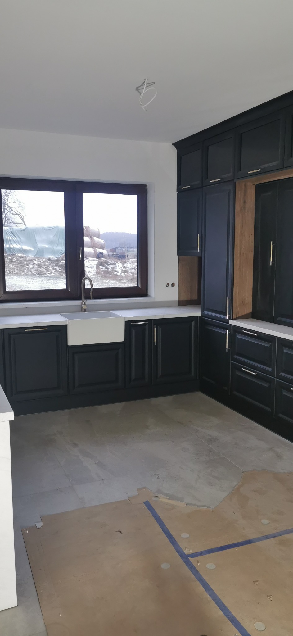 Ciemna zabudowa kuchenna z białym blatem i ceramicznym zlewem farmerskim pod oknem, widok na budowę za oknem, surowy stan podłogi.