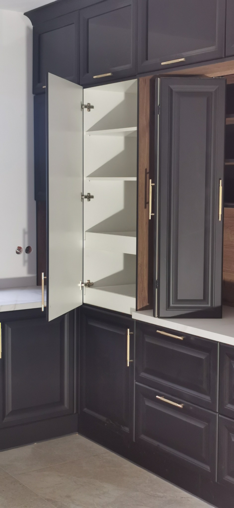 Ciemne szafki kuchenne z białymi półkami wewnątrz, otwarte drzwi ukazują zawiasy, złote uchwyty, widoczne gniazdka w ścianie.