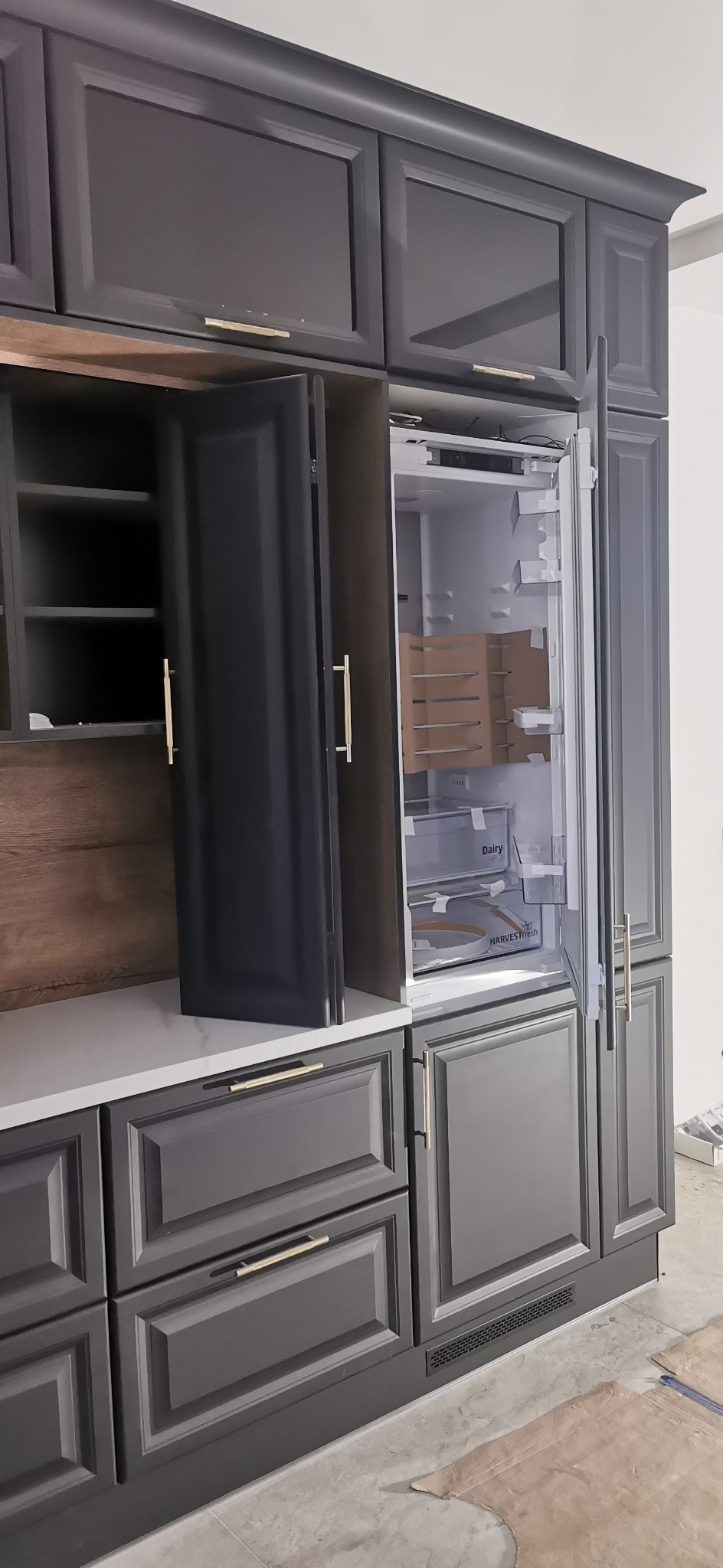 Szara zabudowa kuchenna z otwartymi szafkami i lodówką, złote uchwyty, widoczne elementy wykończenia podłogi.