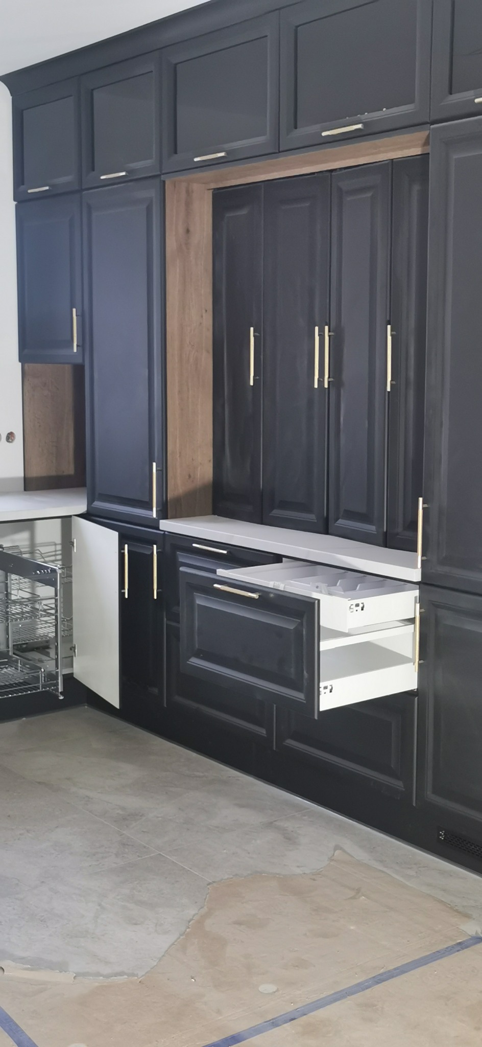 Ciemnoszara zabudowa kuchenna z otwartymi szufladami i koszem cargo, złote uchwyty, widoczny fragment podłogi w trakcie remontu.