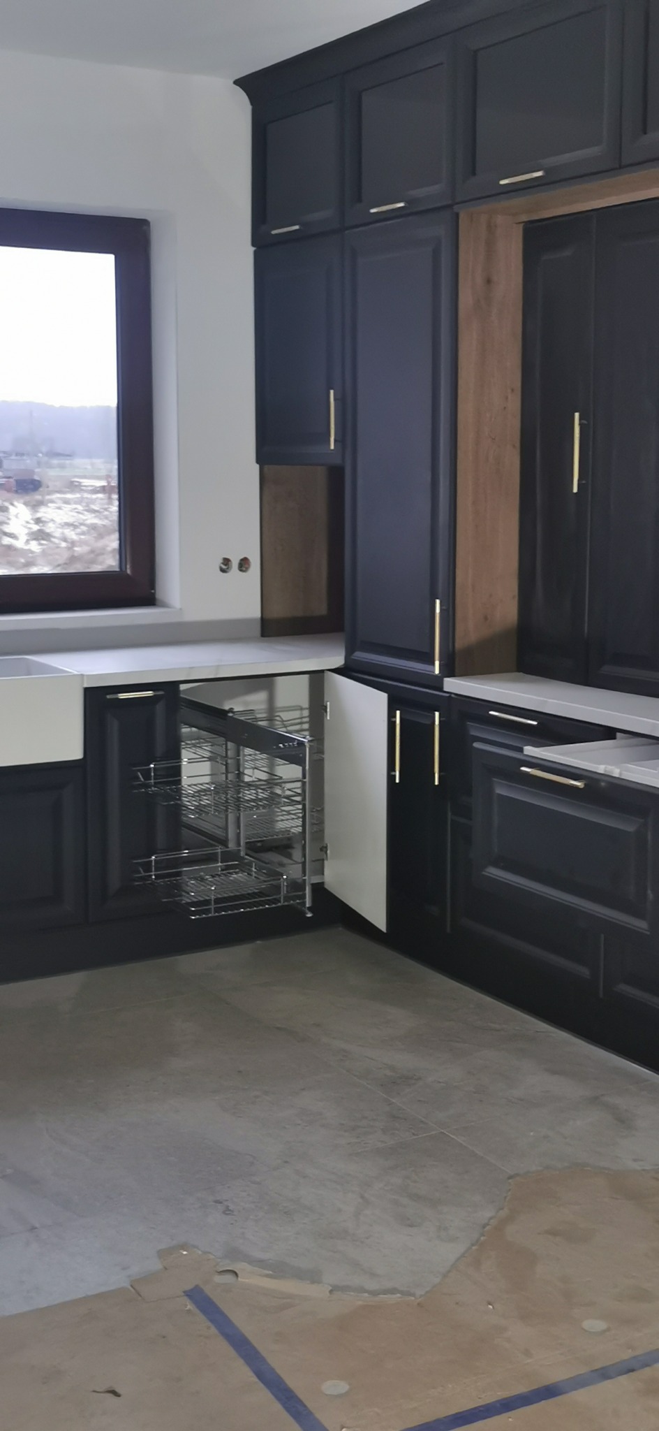 Ciemnogranatowa zabudowa kuchenna z wysuwanym koszem cargo w szafce narożnej, widok na okno i fragment niedokończonej podłogi.