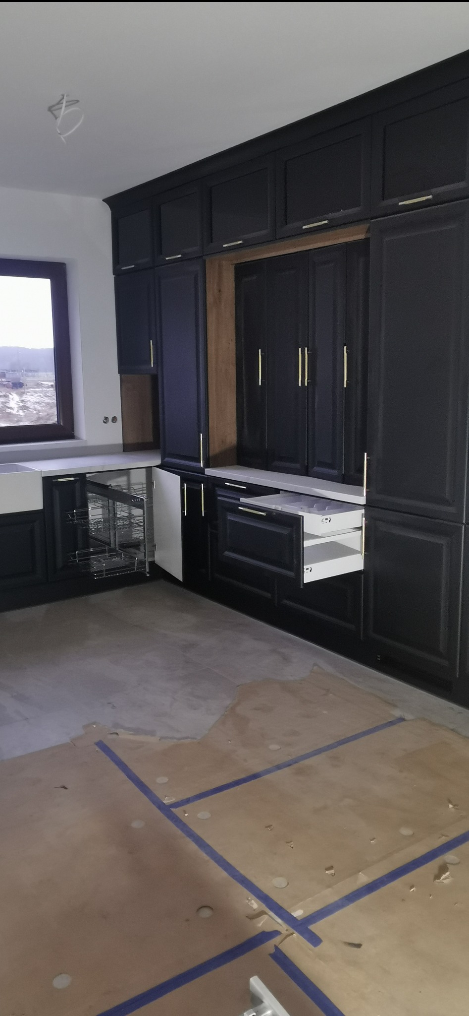 Ciemna zabudowa kuchenna z otwartą szufladą i cargo, złote uchwyty, podłoga zabezpieczona kartonem, widok na krajobraz za oknem.