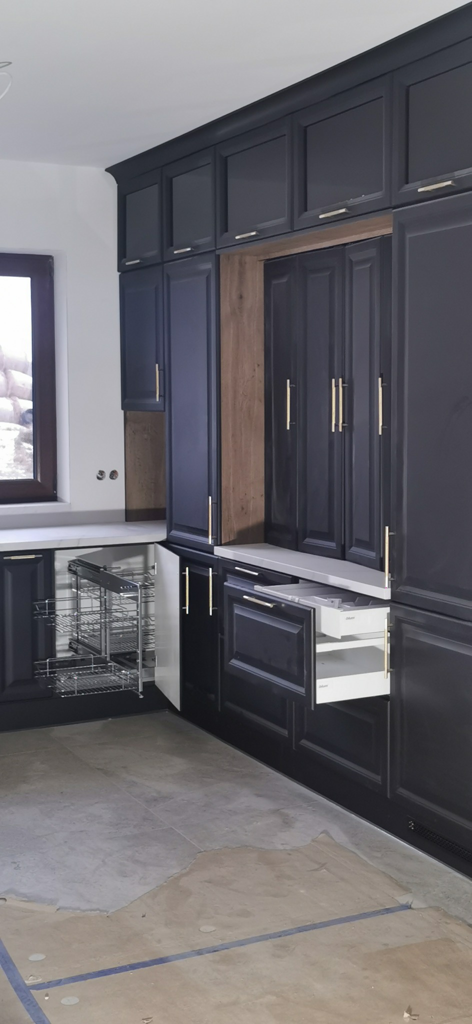 Ciemna zabudowa kuchenna z wysuwanymi szufladami i koszem cargo w rogu, złote uchwyty, surowy stan wykończenia podłogi.