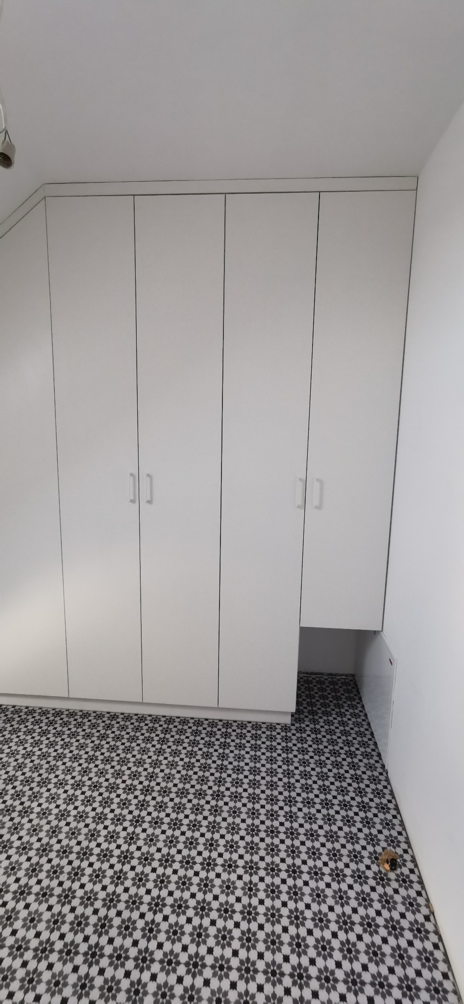 Biała szafa wnękowa z czterema drzwiami, z minimalistycznymi uchwytami, dopasowana do skosu sufitu, na podłodze wzorzyste płytki w odcieniach szarości.