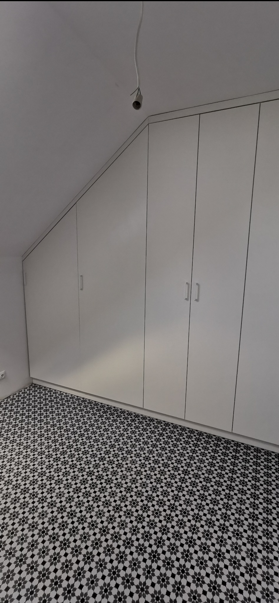 Biała szafa wnękowa pod skosem, z minimalistycznymi uchwytami, na tle wzorzystej podłogi. Widoczny kabel od lampy sufitowej.