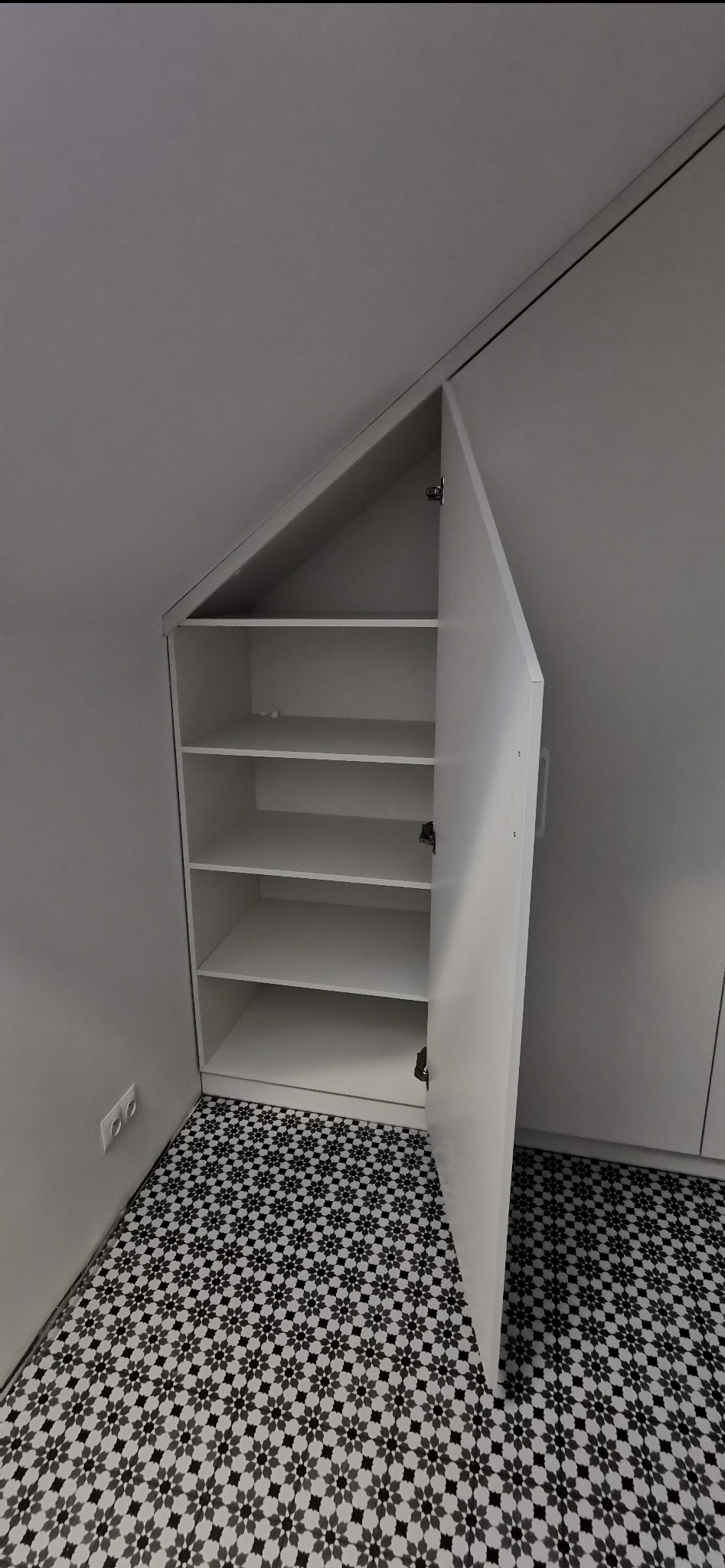 Biała szafa wnękowa z otwartymi drzwiami, dopasowana do skosu dachu, na podłodze wzorzyste płytki. Widoczne półki wewnątrz szafy.