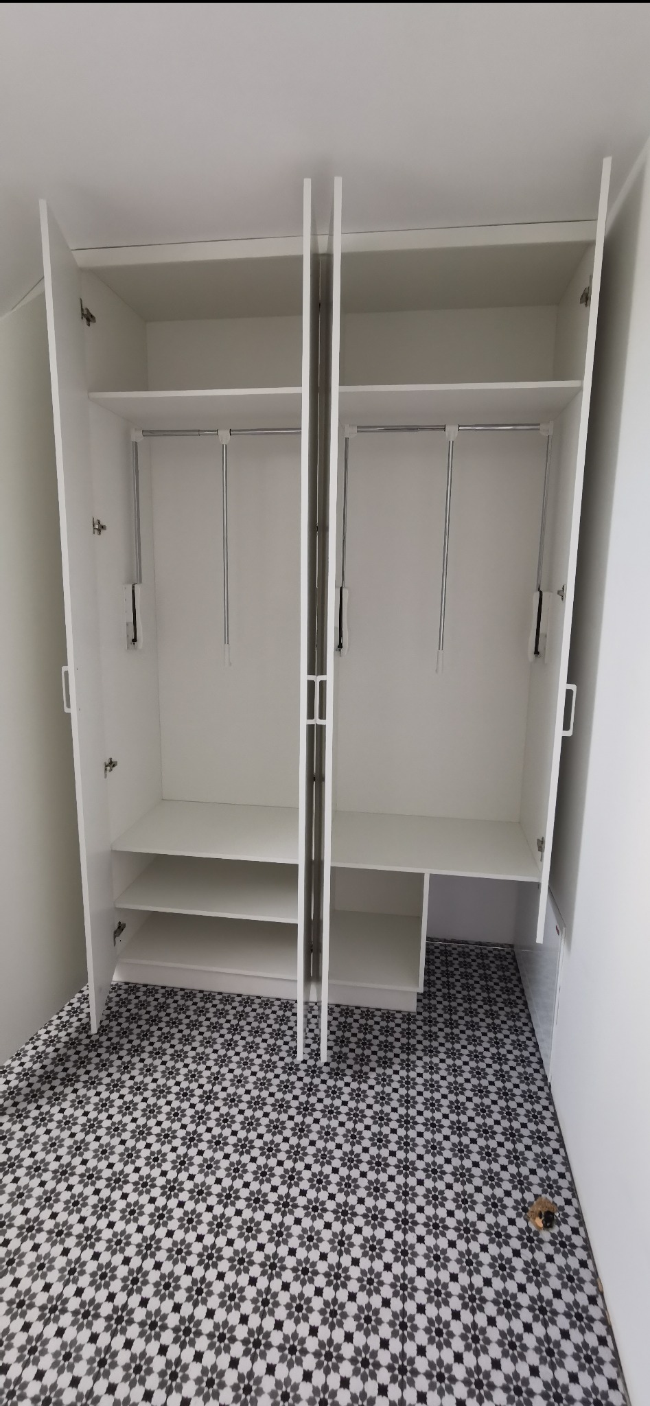 Biała szafa wnękowa z otwartymi drzwiami, widoczne półki i drążki na ubrania. Na podłodze wzorzyste płytki. Minimalistyczny design.