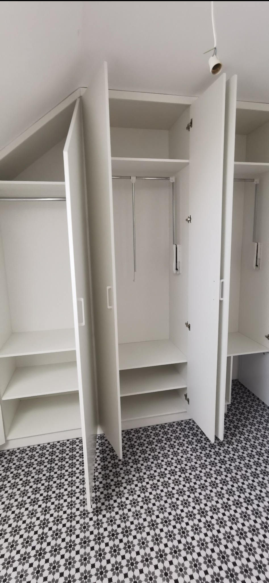 Biała szafa wnękowa z otwartymi drzwiami, widoczne półki i drążki na ubrania, podłoga w czarno-białe wzory. Szafa dopasowana do skosu dachu.