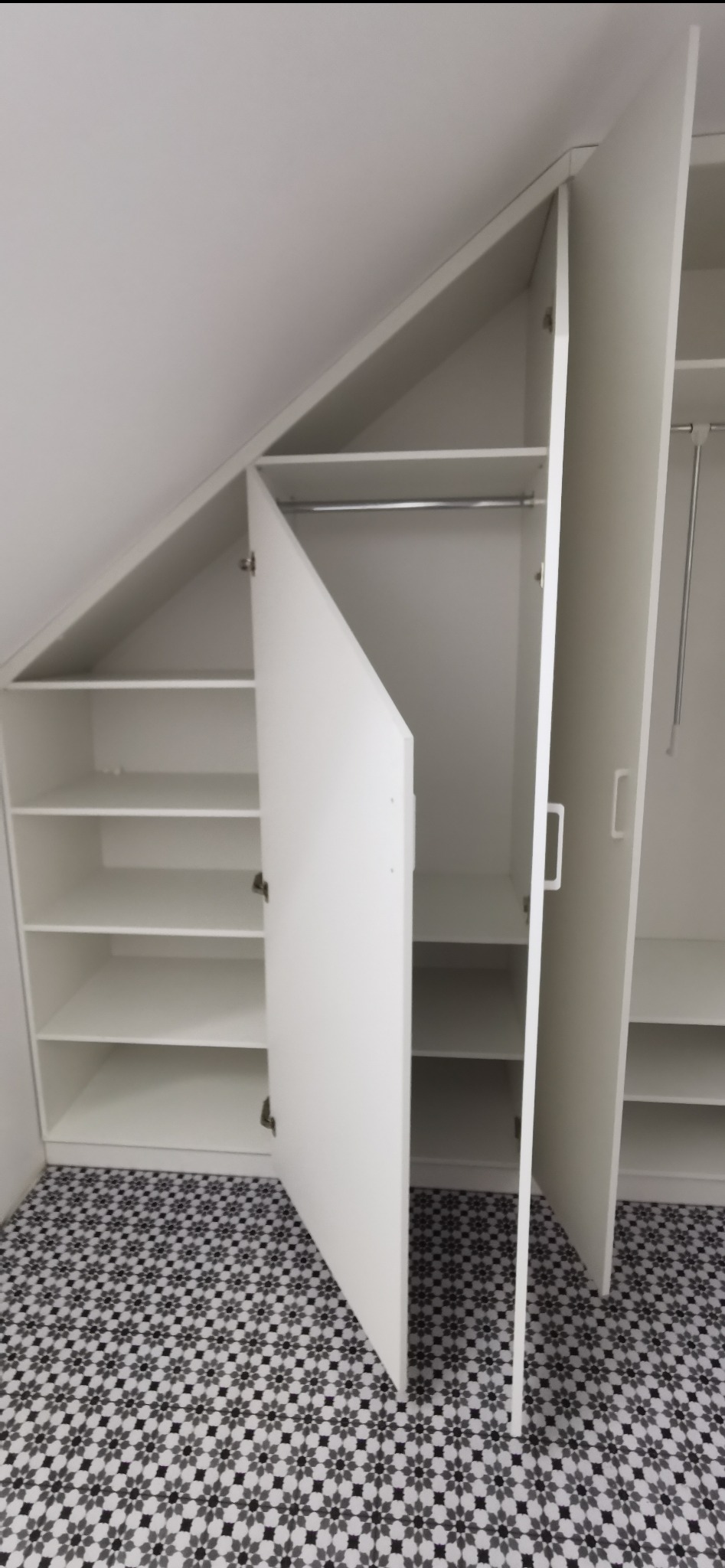 Biała szafa wnękowa z otwartymi drzwiami, dopasowana do skosu dachu, na podłodze wzorzyste płytki. Widoczne półki i drążek na ubrania.