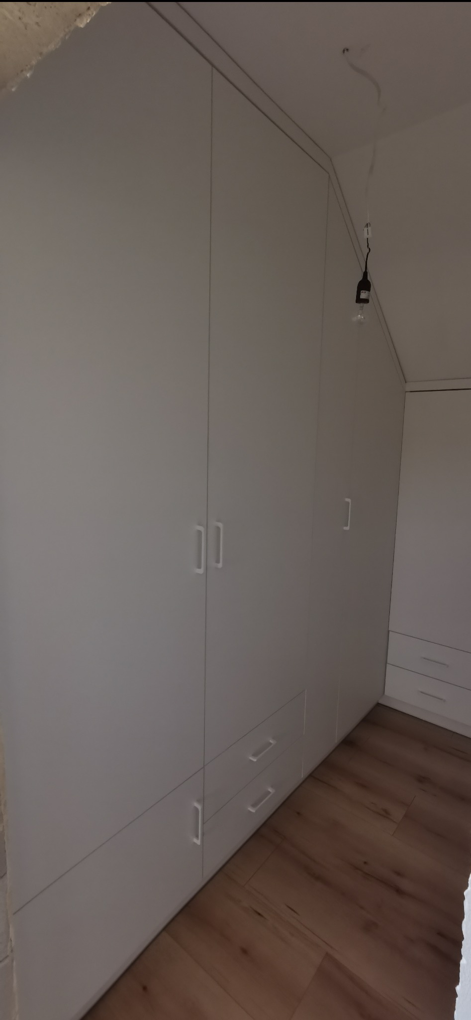 Biała szafa wnękowa z szufladami i minimalistycznymi uchwytami, dopasowana do skosu dachu, na tle drewnianej podłogi. Widoczny kabel oświetleniowy.