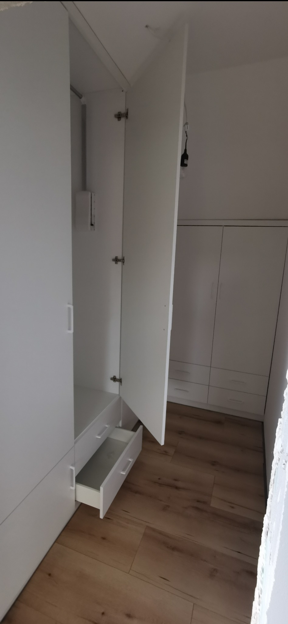 Biała szafa wnękowa z otwartymi drzwiami i wysuniętą szufladą, widok na wnętrze szafy i podłogę z paneli. W tle kolejna szafa w zabudowie.