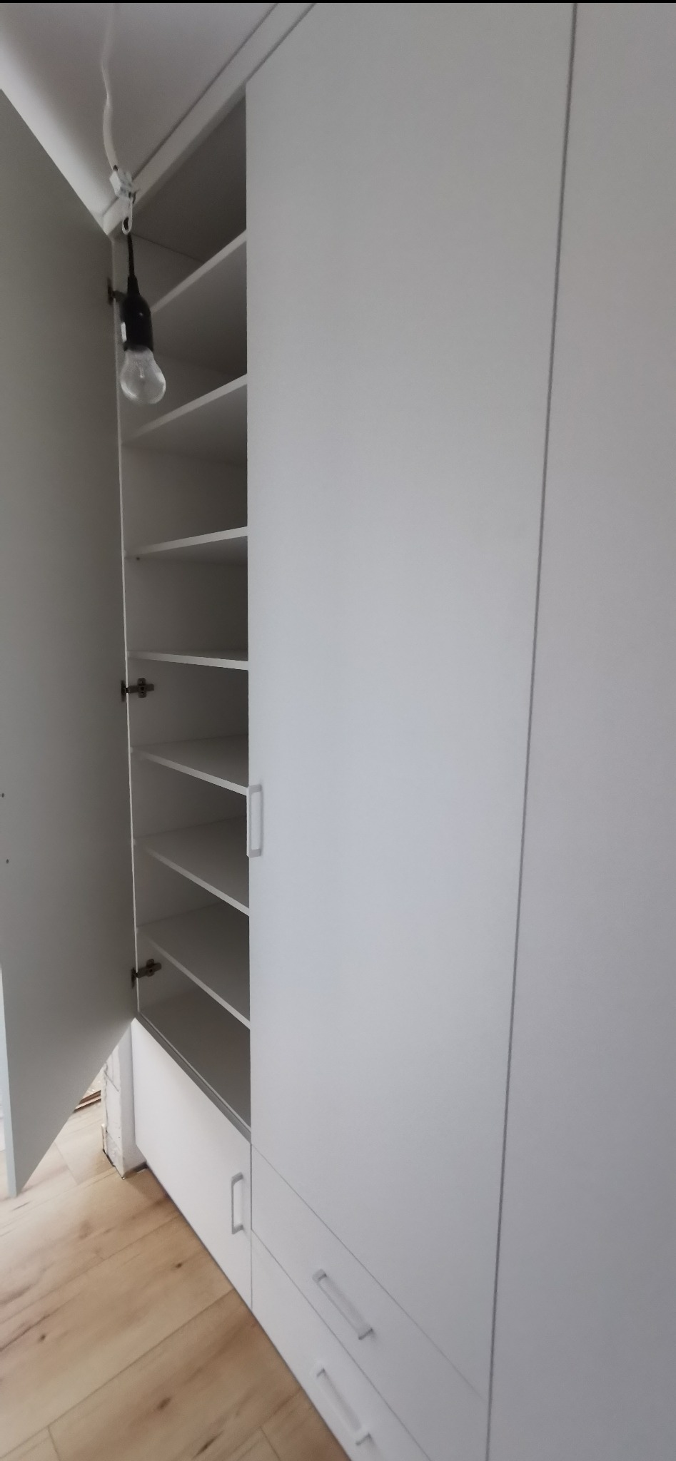 Biała szafa wnękowa z otwartymi drzwiami, ukazująca półki. Minimalistyczny design, widoczne szuflady na dole. Uwagę zwraca prowizoryczne oświetlenie wewnątrz szafy.