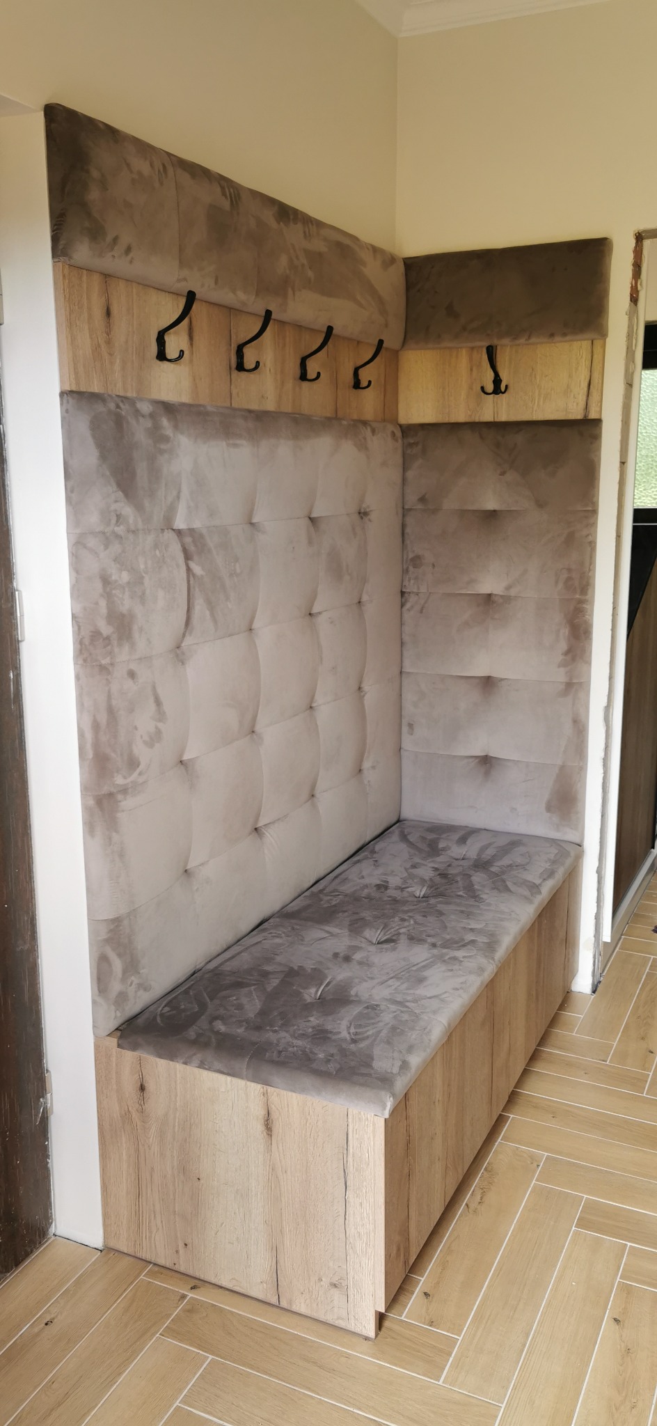 Kącik w przedpokoju z siedziskiem i wieszakami, obity beżową tapicerką z pikowaniem. Drewnopodobne wykończenie, czarne haczyki na ubrania.