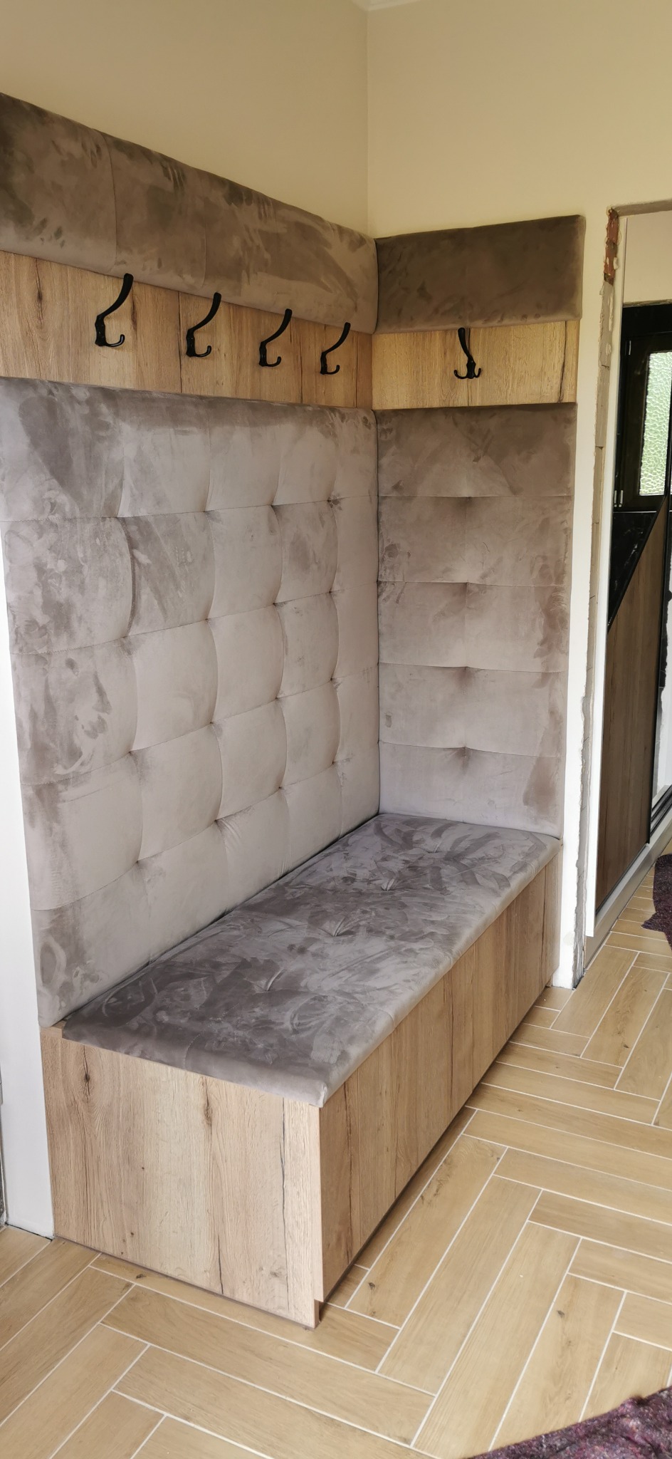 Tapicerowany narożnik z wieszakami w przedpokoju. Drewnopodobne wykończenie, pikowane oparcie i siedzisko w odcieniach szarości. Funkcjonalne rozwiązanie do przechowywania odzieży.