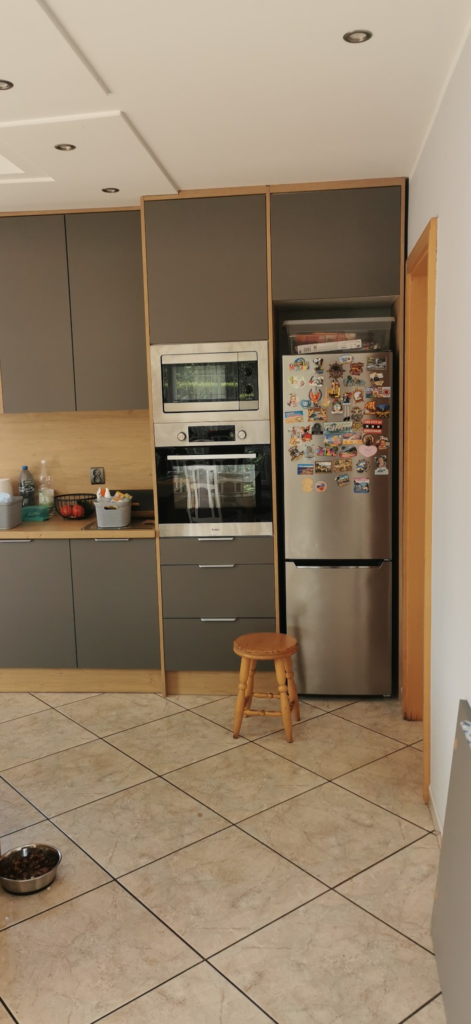 Szara zabudowa kuchenna z drewnianymi akcentami, piekarnikiem, mikrofalówką i lodówką. Na podłodze stoi drewniany taboret. Widoczna miska dla zwierząt.
