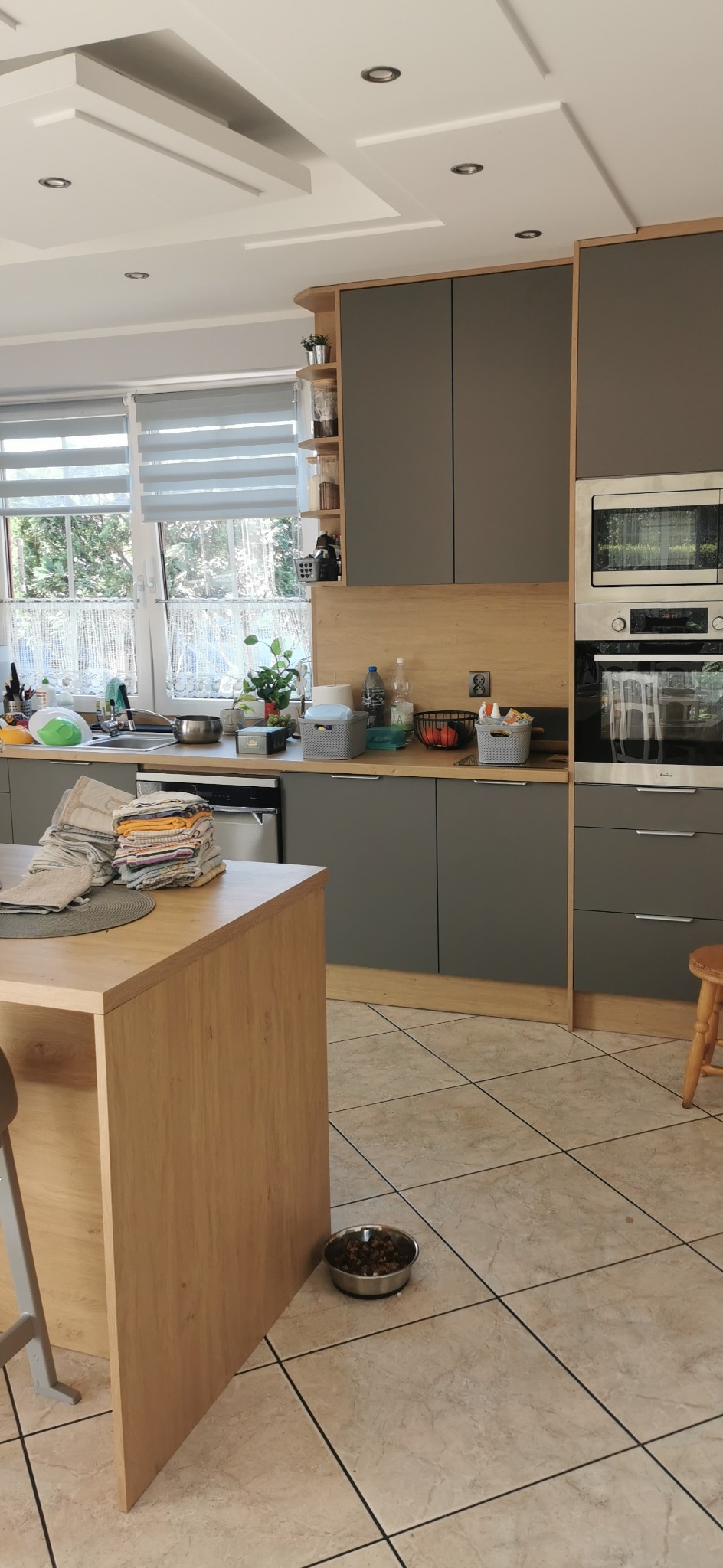 Szara zabudowa kuchenna z drewnianymi akcentami, wyspa kuchenna z blatem i stosem ścierek, okno z roletami w tle. Miska z karmą dla zwierząt na podłodze.