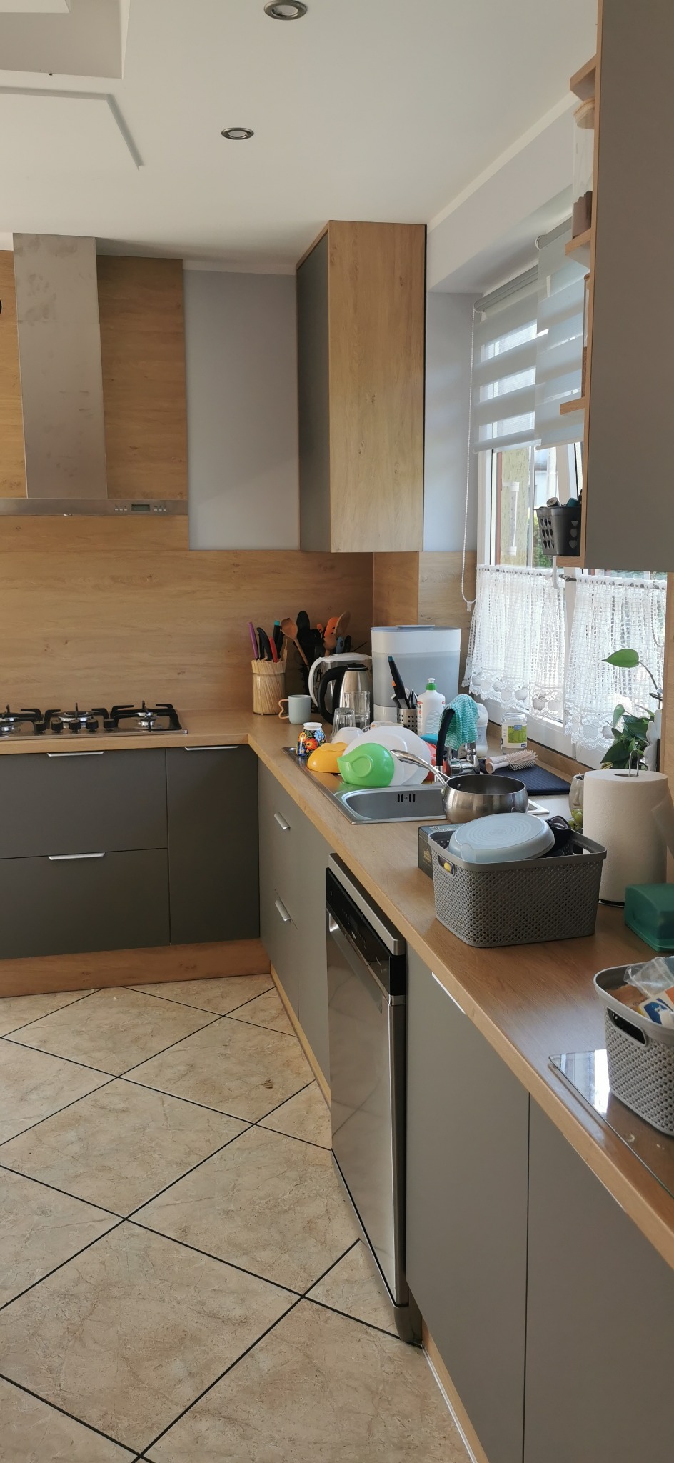 Szare szafki kuchenne z drewnianymi blatami i sprzętem AGD. Widoczny fragment płyty grzewczej i zlewu z naczyniami. Ujęcie z perspektywy podłogi, z widoczną mozaiką.