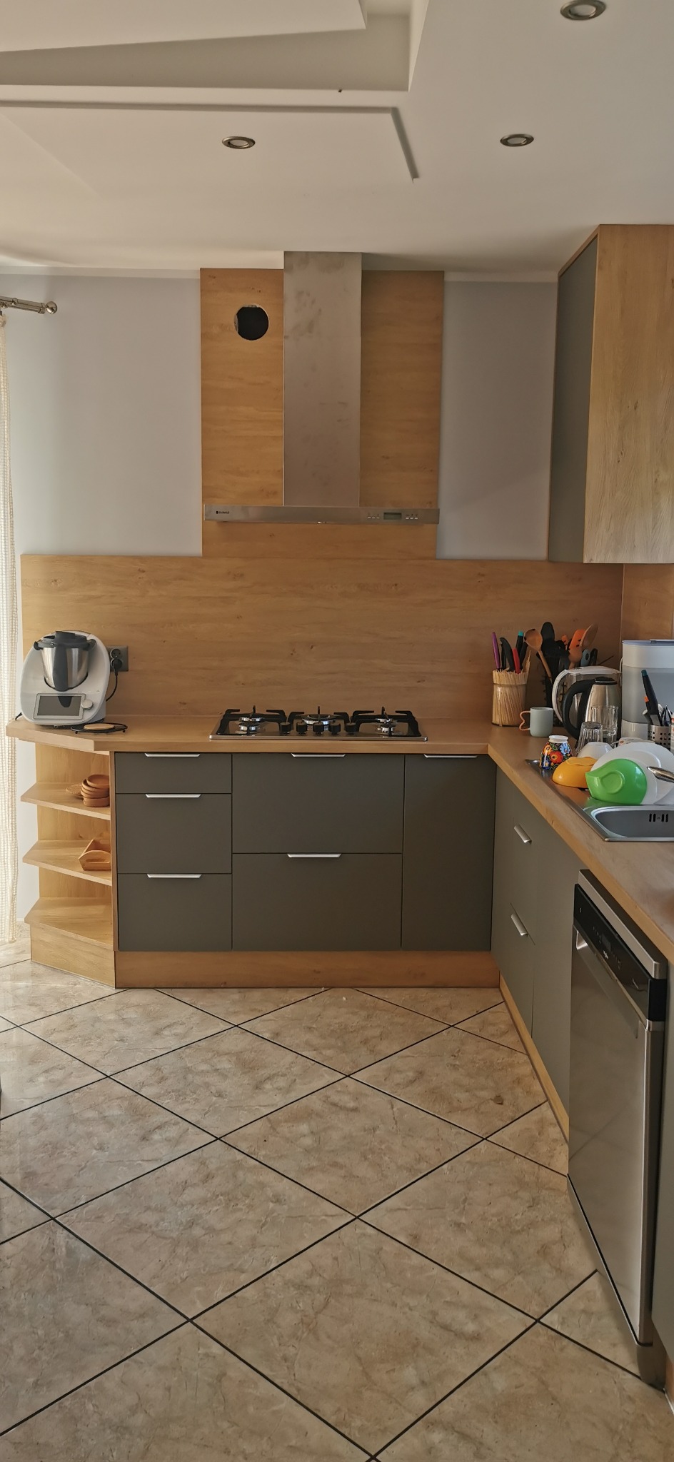 Nowoczesna kuchnia w odcieniach szarości i drewna z blatem roboczym, płytą gazową i okapem. Widoczny sprzęt AGD i akcesoria kuchenne. Ujęcie z perspektywy podłogi.