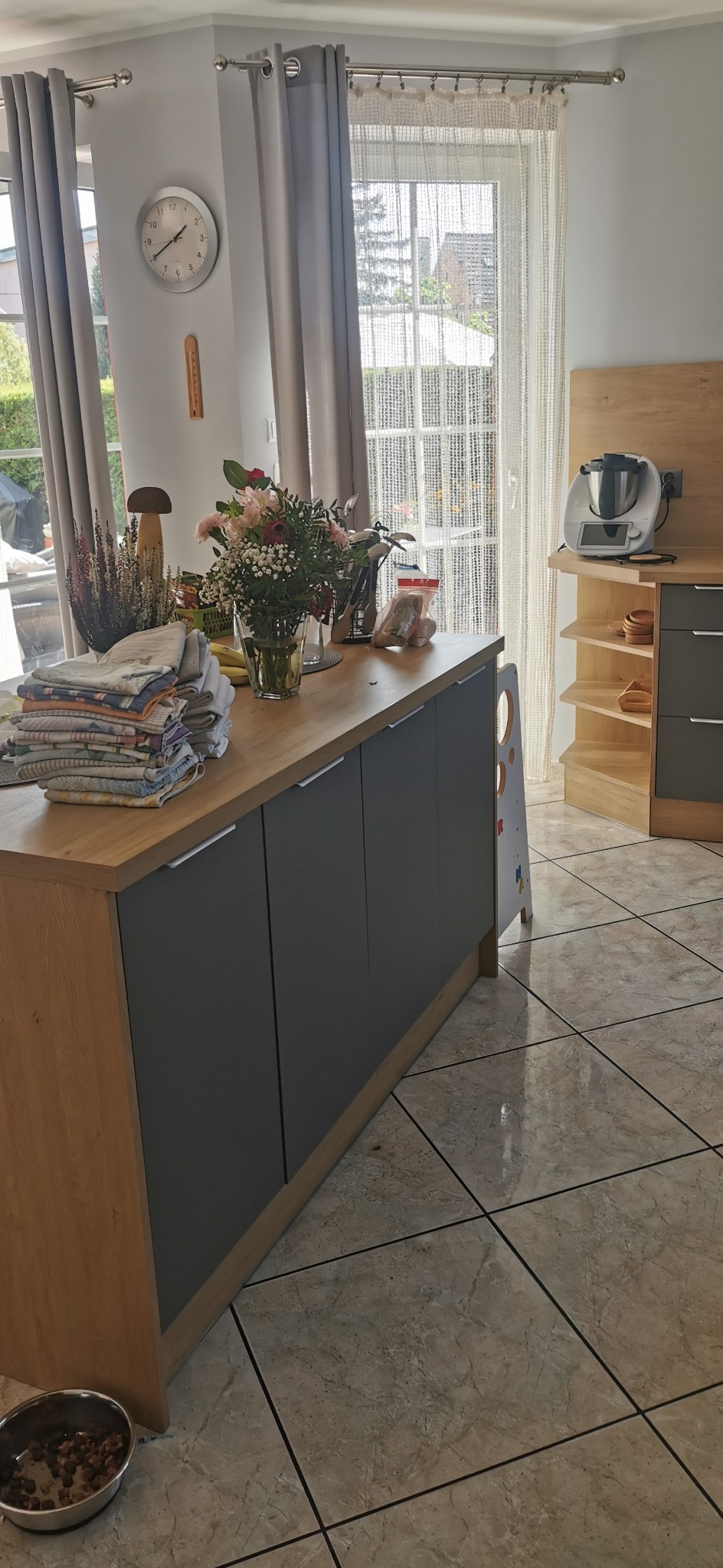 Jasna kuchnia z wyspą i szarymi frontami. Na blacie kwiaty, naczynia i tekstylia. W tle okno z firanką i sprzęt AGD. Na podłodze płytki. Widok na wnętrze domu.