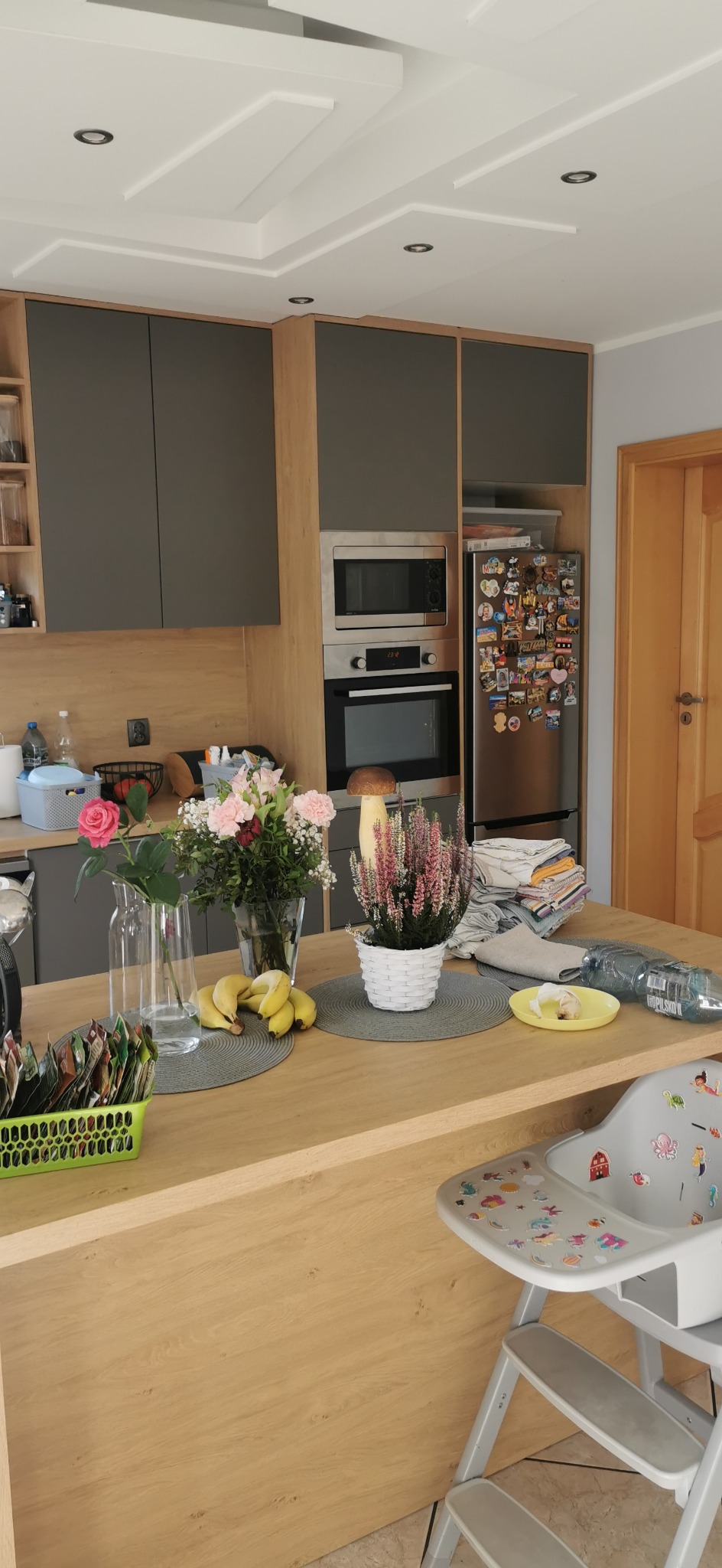 Nowoczesna kuchnia z drewnianym blatem wyspowym, szarymi szafkami i sprzętem AGD. Na blacie kwiaty, owoce i dziecięce krzesełko ze naklejkami.
