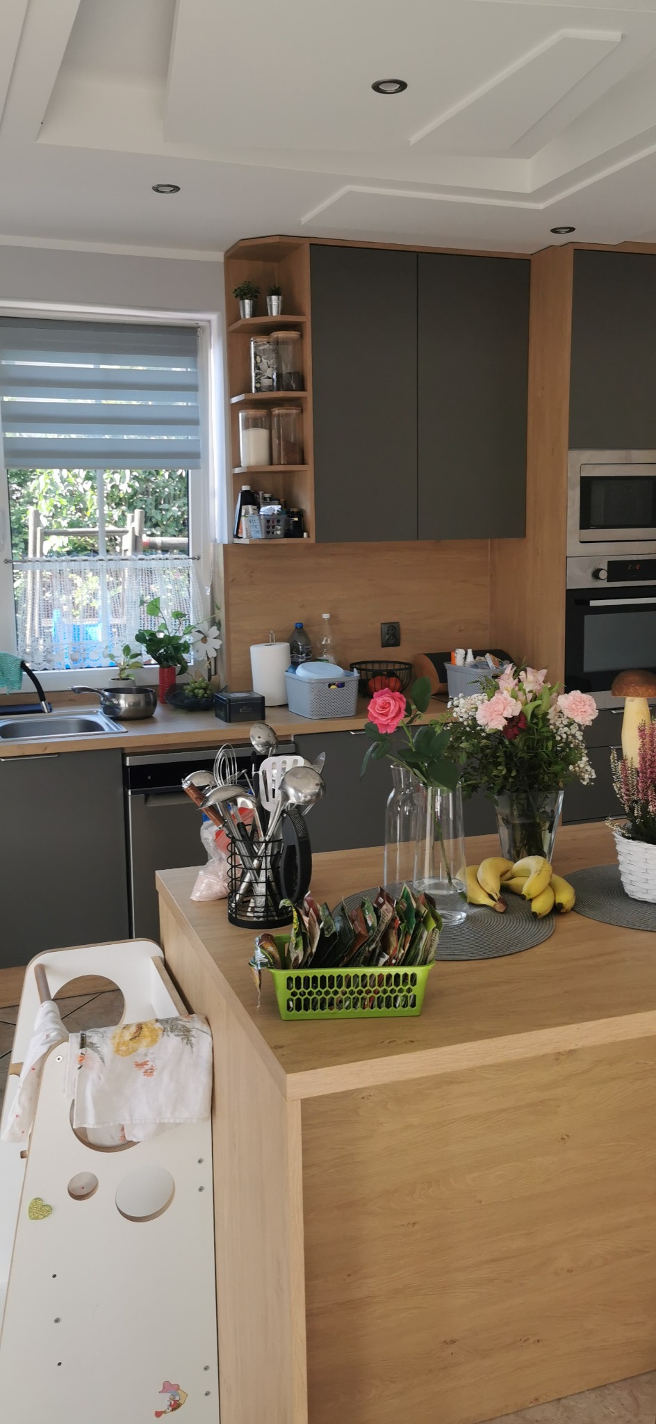 Nowoczesna kuchnia z szarymi szafkami i drewnianym blatem. Na blacie kwiaty, owoce i akcesoria kuchenne. Widoczne okno z roletami i krzesełko dla dziecka.