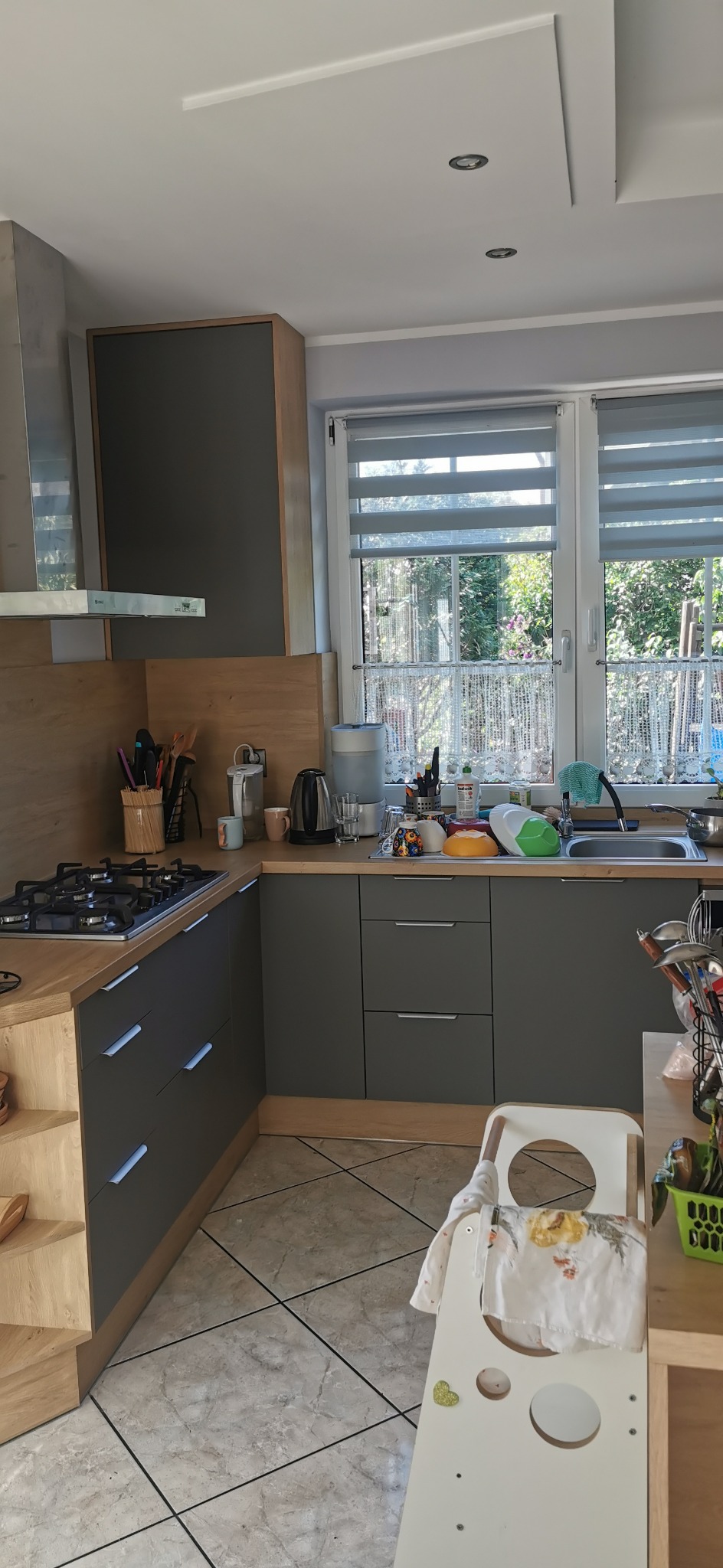 Szara kuchnia w kształcie litery L z drewnianymi blatami i srebrnymi uchwytami. Widoczne okno z roletami i dziecięce krzesełko. Umyte naczynia w zlewie.