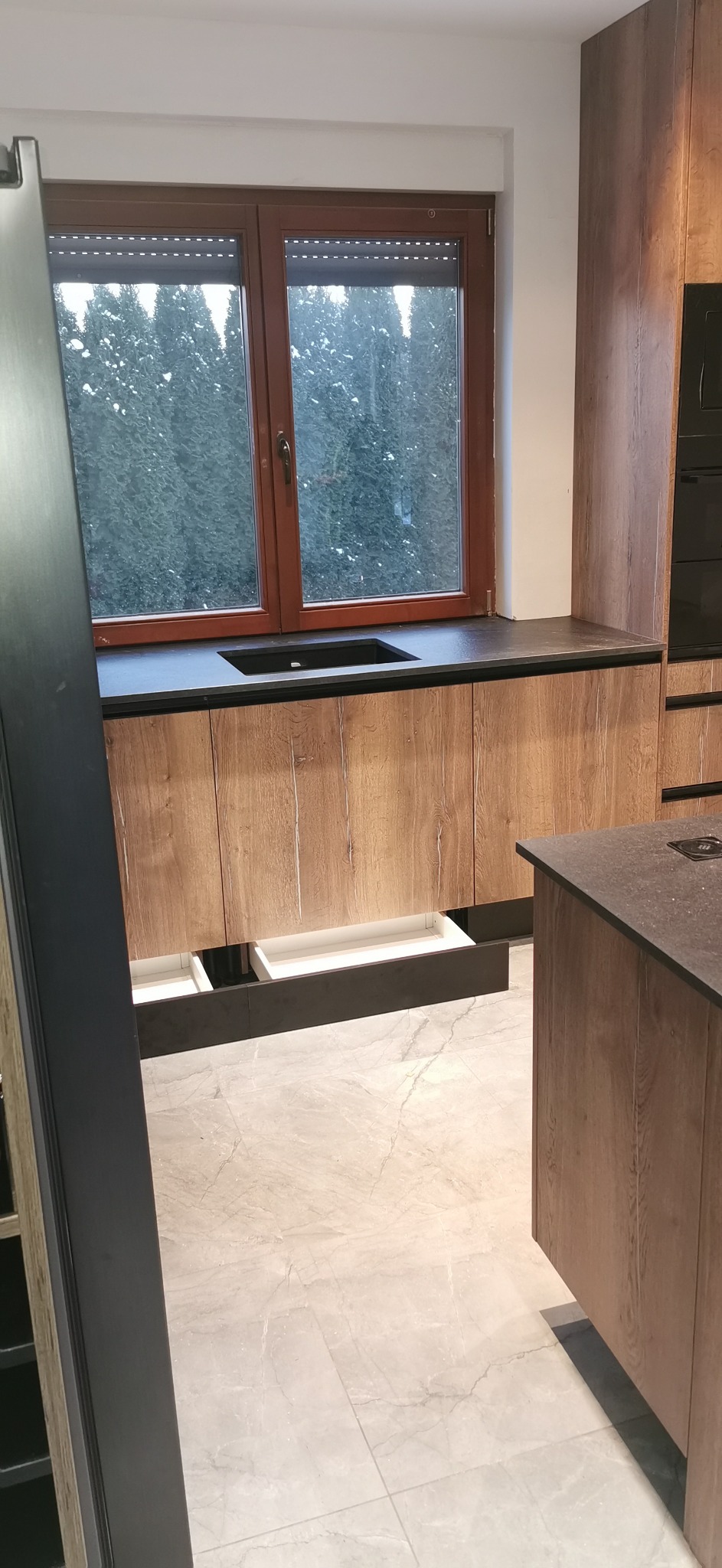 Nowoczesna kuchnia z drewnianymi frontami i czarnym blatem pod oknem. Wysuwane szuflady, marmurowa podłoga. Widok na zewnątrz przez okno.