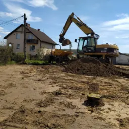 Żółta koparka nabiera ziemię i wysypuje ją do wywrotki w Królówce, w tle dom jednorodzinny i błękitne niebo z chmurami.