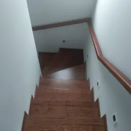 Drewniane schody z brązową balustradą w jasnym wnętrzu, widok z góry w kierunku okna dachowego.