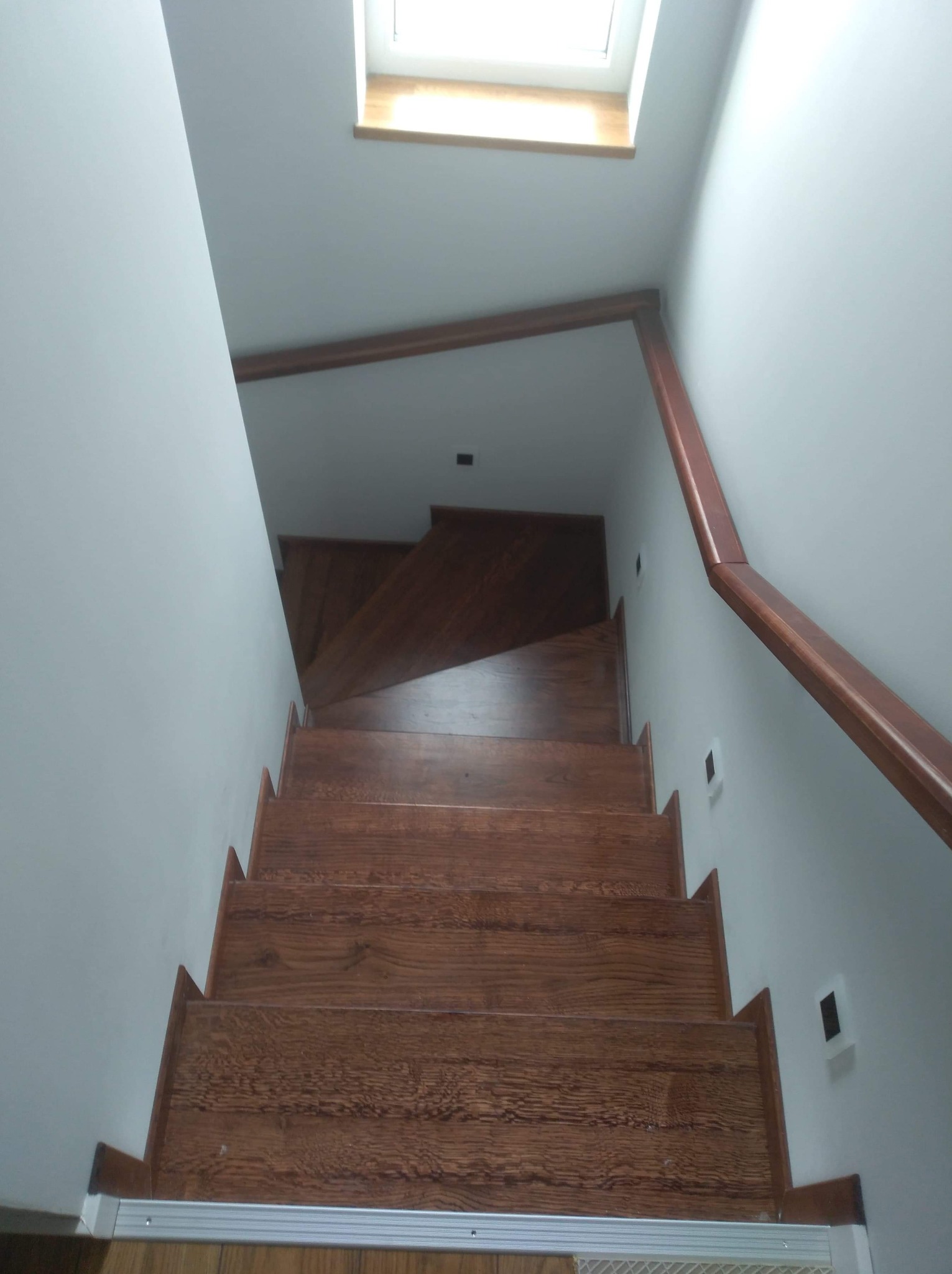 Drewniane schody z brązową balustradą w jasnym wnętrzu, widok z góry w kierunku okna dachowego.