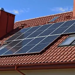 Panele słoneczne zamontowane na dachu z dachówką ceramiczną, widoczne okna dachowe i komin.