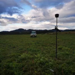 Pomiar geodezyjny w terenie otwartym: samochód terenowy i sprzęt pomiarowy na łące pod pochmurnym niebem.