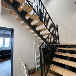 STALIAS Krystian Białas - Nowoczesne schody z drewnianymi stopniami i czarną metalową konstrukcją, widok z dołu, minimalistyczne wnętrze z białymi ścianami i drewnianą podłogą, barierka zabezpieczająca dla dzieci na dole...