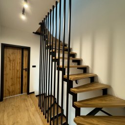 STALIAS Krystian Białas - Nowoczesne schody drewniane z czarną, pionową balustradą, oświetlenie punktowe sufitowe, widok z dołu na górę, drzwi w tle.