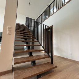 STALIAS Krystian Białas - Nowoczesne schody metalowe z drewnianymi stopniami i czarną balustradą, widziane z dołu, z widocznymi lampami wiszącymi i fragmentem balustrady na antresoli.