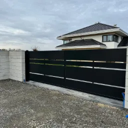 Nowoczesna, przesuwna brama wjazdowa w kolorze czarnym z poziomymi przerwami, zamontowana między betonowymi słupkami na tle dwupiętrowego domu z ciemną dachówką i pochmurnym niebem.